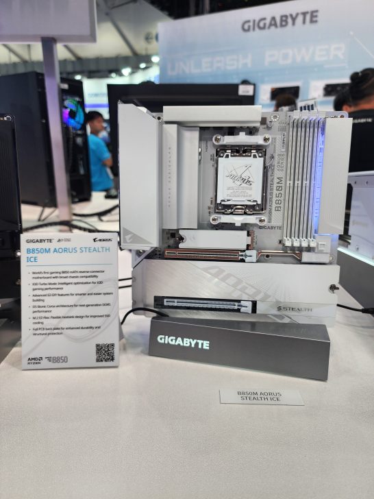 The image shows a Gigabyte B650M AORUS Stealth ICE motherboard displayed on a stand with signage highlighting its features at a tech event.