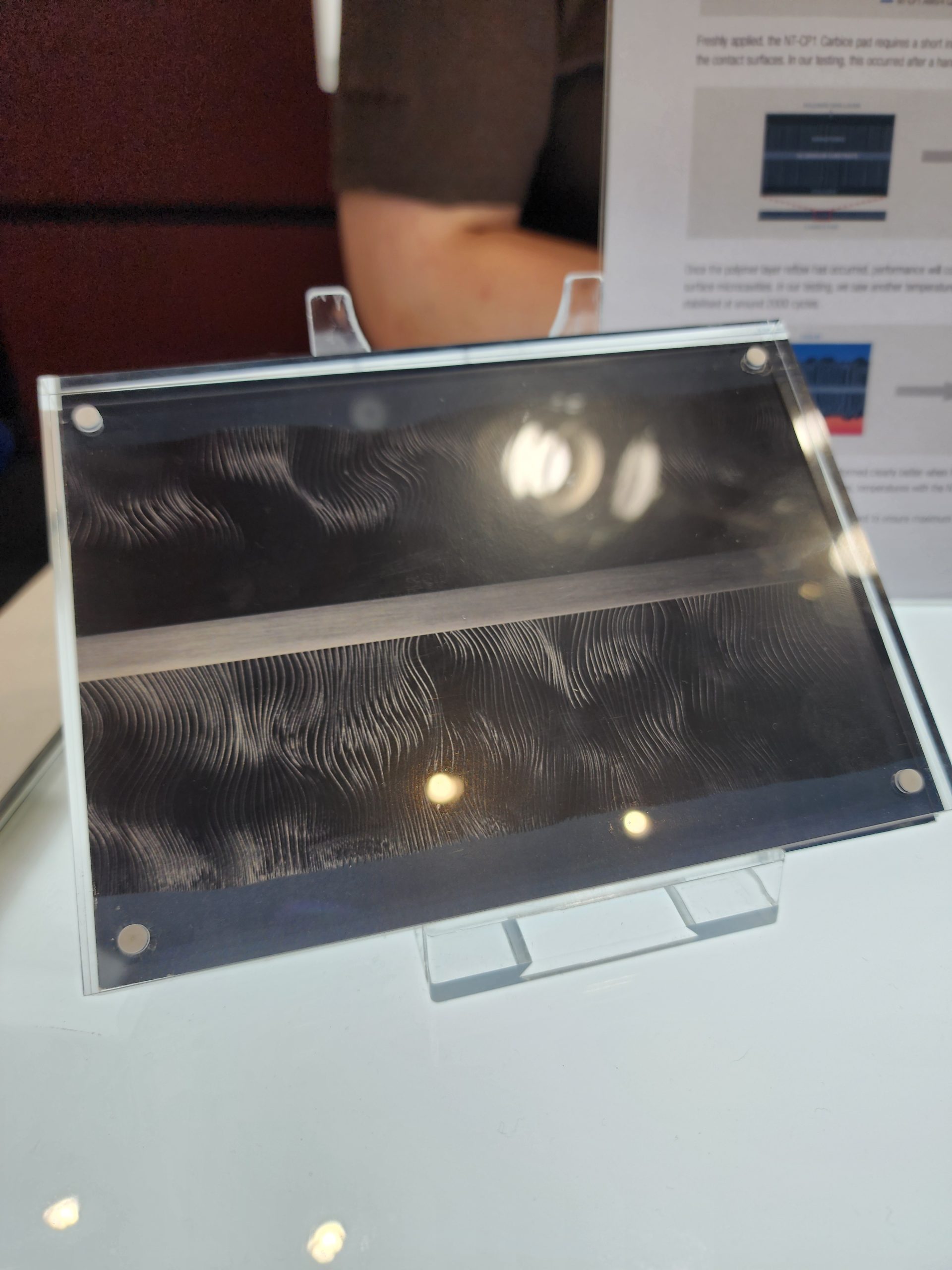 A close-up of a carbon pad with a wave-like texture displayed on a transparent stand, with partial text from a technical document visible in the background.