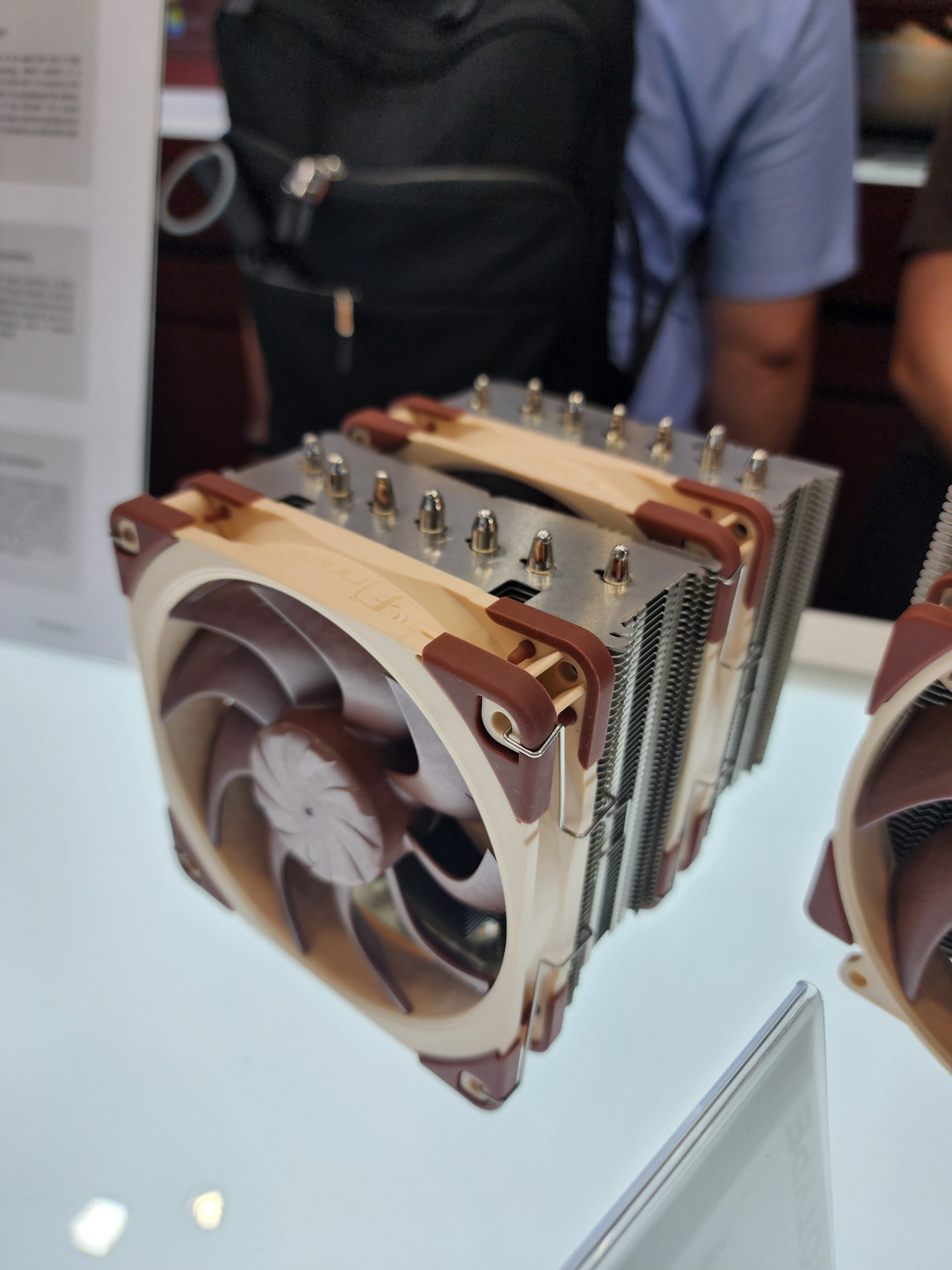 A Noctua dual-tower CPU cooler with two fans displayed, surrounded by people.
