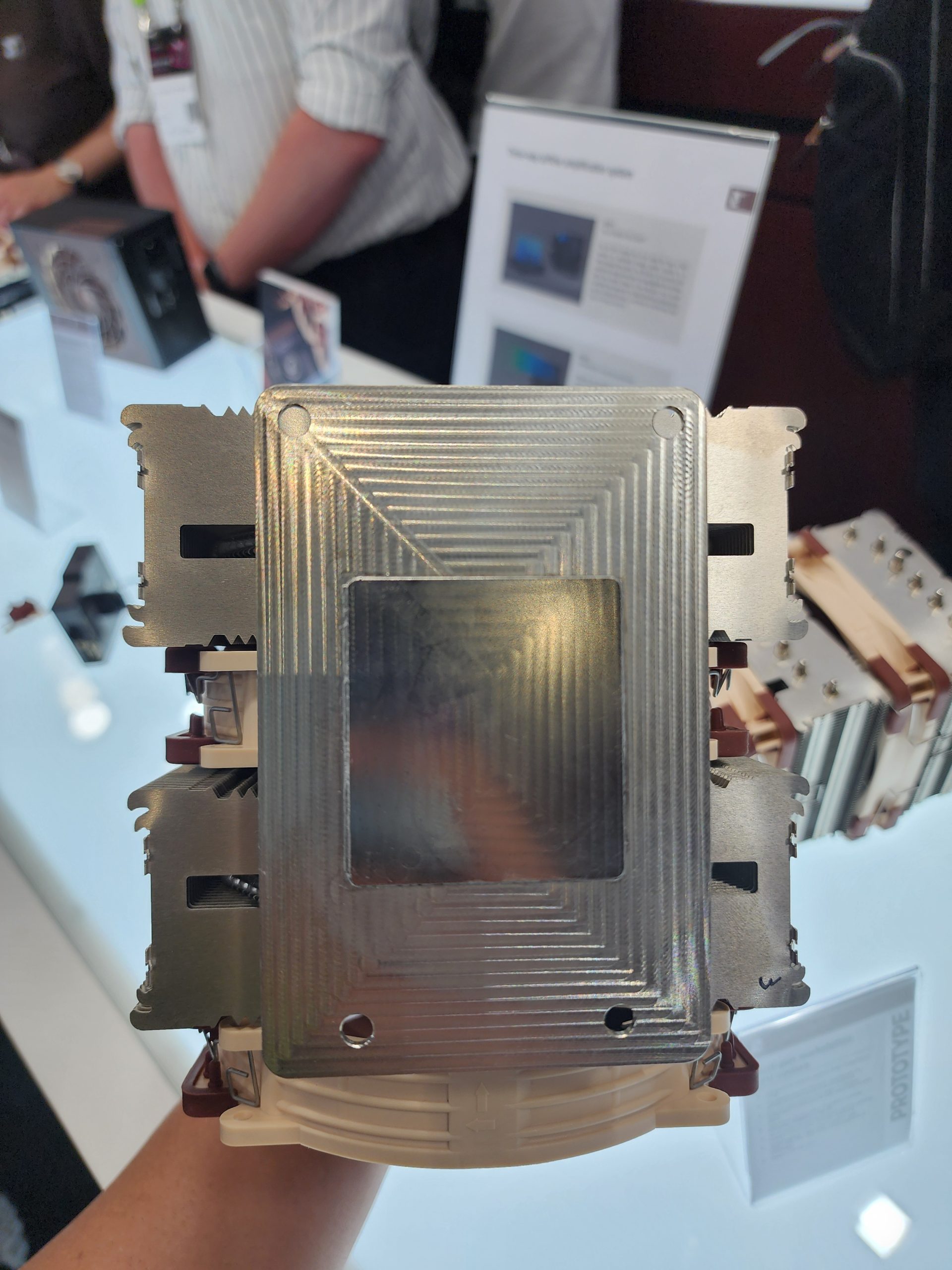 A person holds a large CPU cooler prototype with multiple metal fins and a reflective top cover, displayed at a tech event booth.