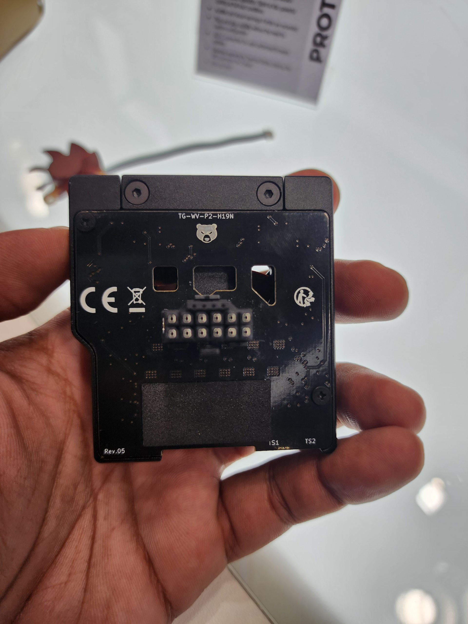 A person holds a component labeled 'TG-WV-P2-H19N' and 'Rev.05', with visible connectors and a partially obscured display stand in the background.