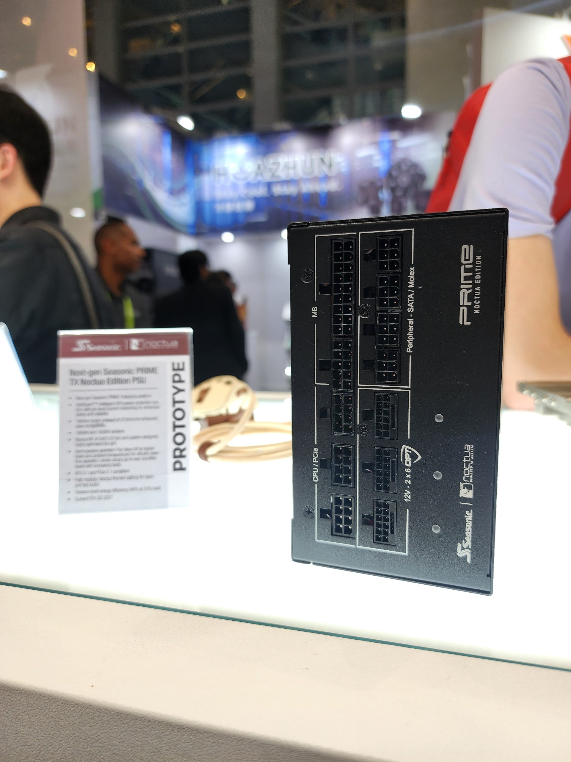 A Seasonic PRIME power supply prototype labeled '12V 24-pin ATX' and 'NEXT-GEN' on display at a tech convention booth, accompanied by a sign describing its features and collaboration with Noctua.