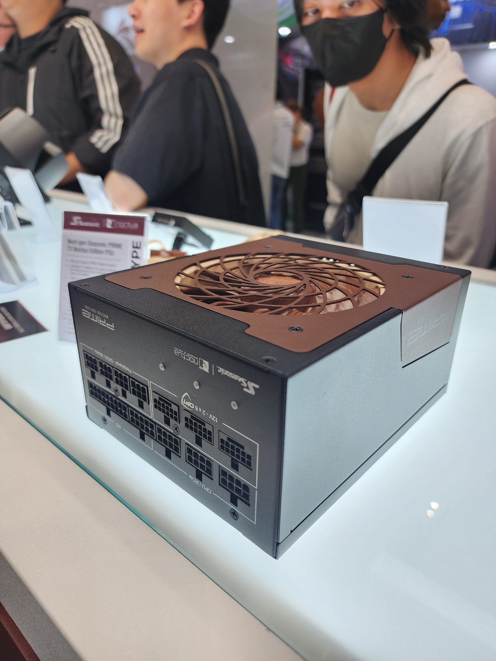 A Seasonic PRIME TX Noctua Edition PSU is displayed on a counter, surrounded by people at an exhibition.