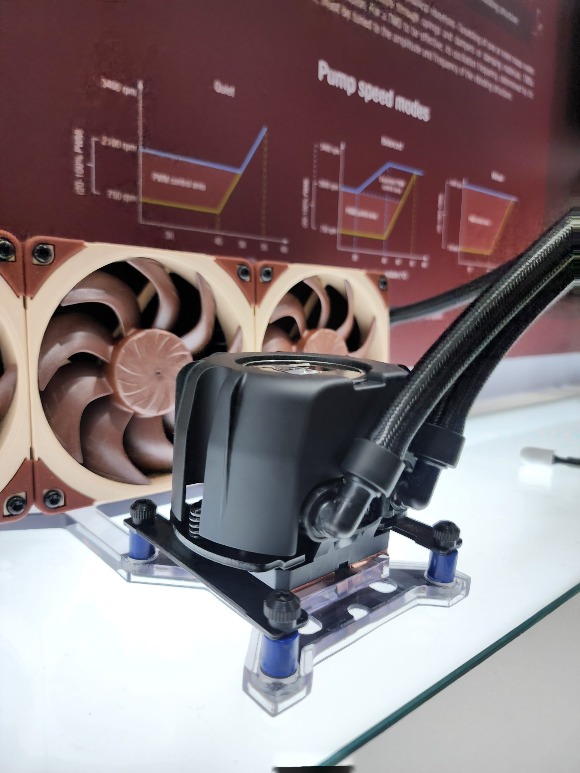 A close-up of a PC cooling solution with brown Noctua fans and tubes, in front of a display with pump speed modes graph labeled 'Quiet' and 'Ultra Quiet.'