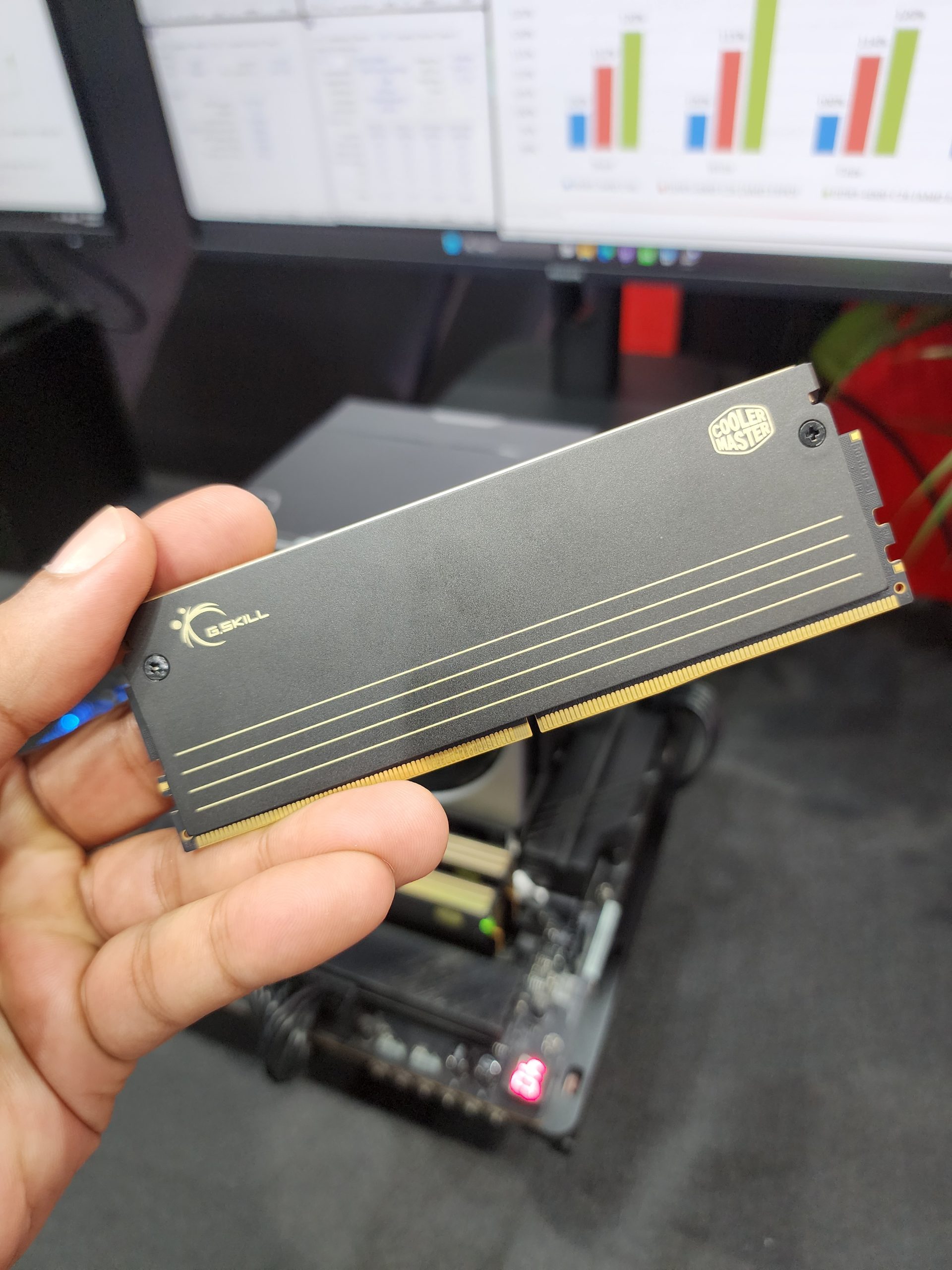 A person holds a G.SKILL RAM module with a Cooler Master label in front of benchmarking software displayed on computer monitors.