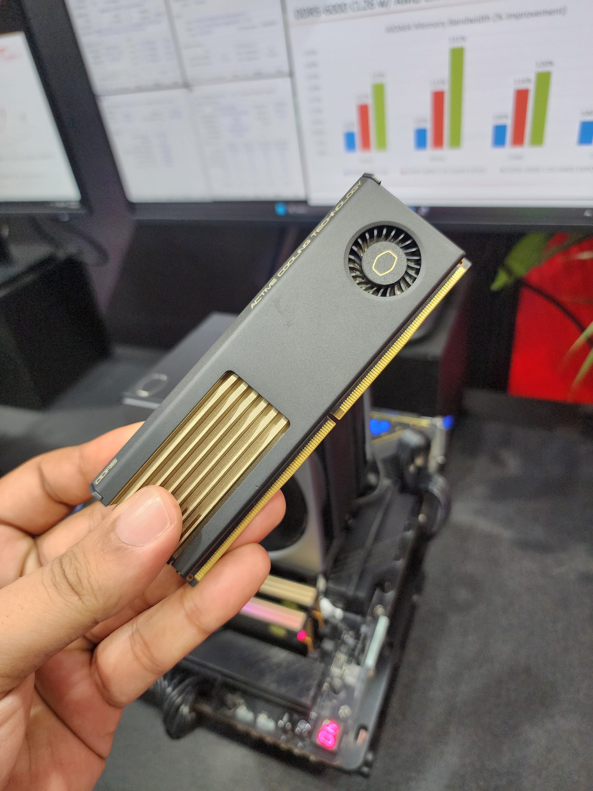 A person holds a Cooler Master active cooling module in front of computer monitors displaying benchmark charts and data.