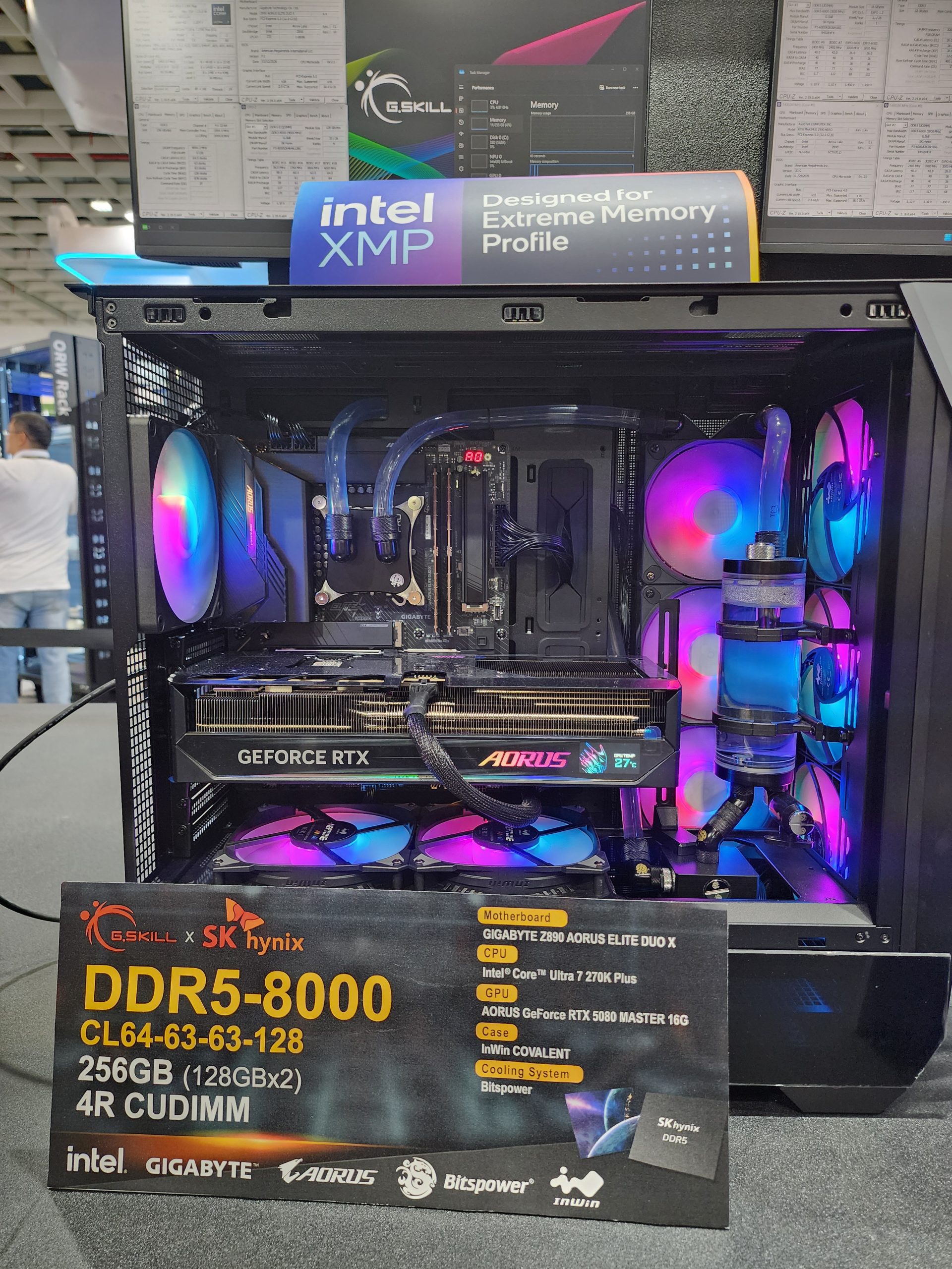 A high-end custom PC setup is displayed featuring a G.SKILL DDR5-8000 RAM and an AORUS GeForce RTX 5080 Master 16G GPU, with signage highlighting Intel's XMP support and a list of components including 'Intel Core Ultra 7 270K Plus' CPU and Gigabyte Z690 AORUS Elite Duo X motherboard.