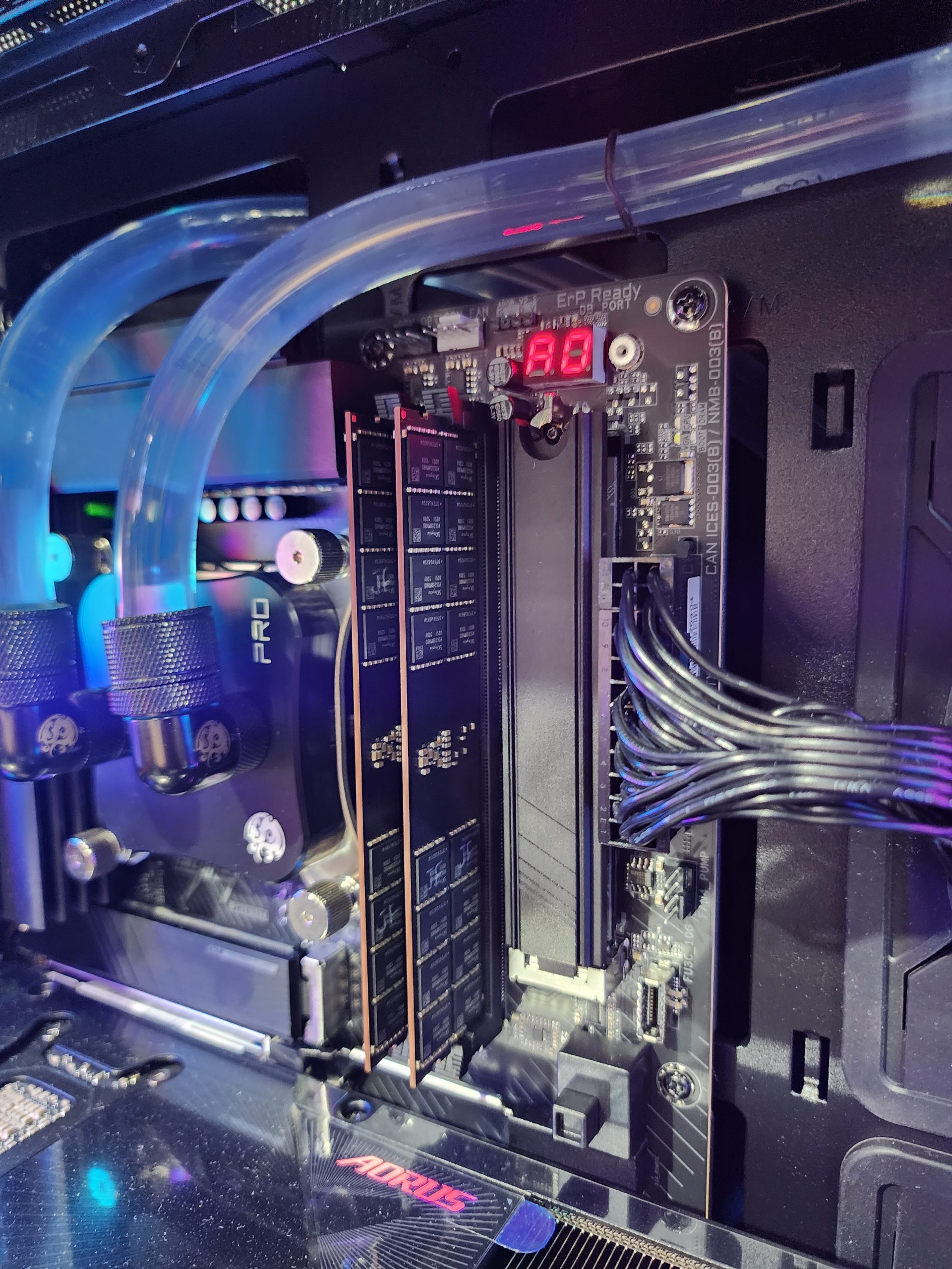 A custom PC build featuring an AORUS motherboard with visible illuminated tubing, adjacent to a digital display showing '98' and a set of installed RAM modules.