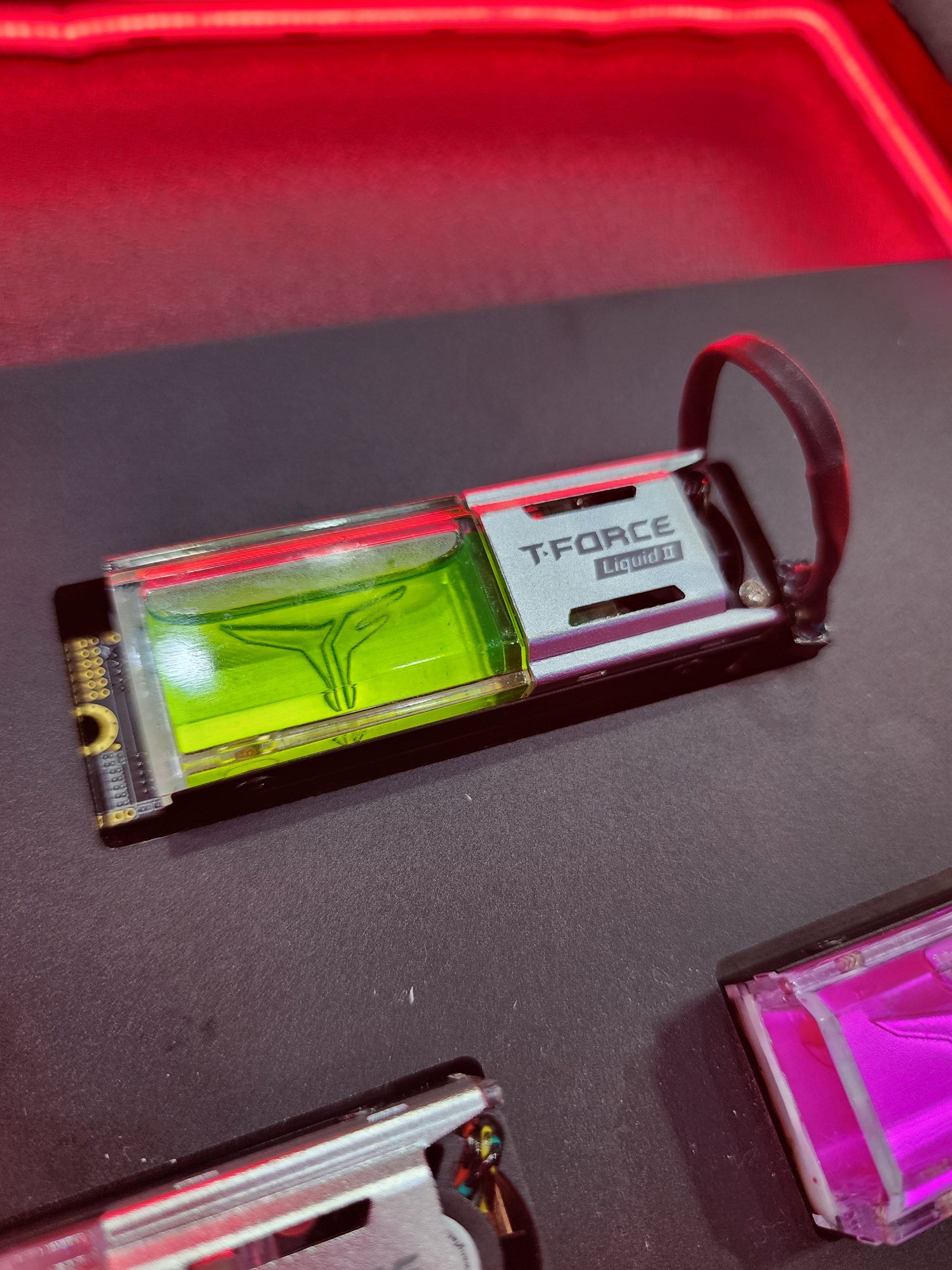 A T-Force Liquid II SSD with a visible green liquid cooling module, set against a red-lit background.