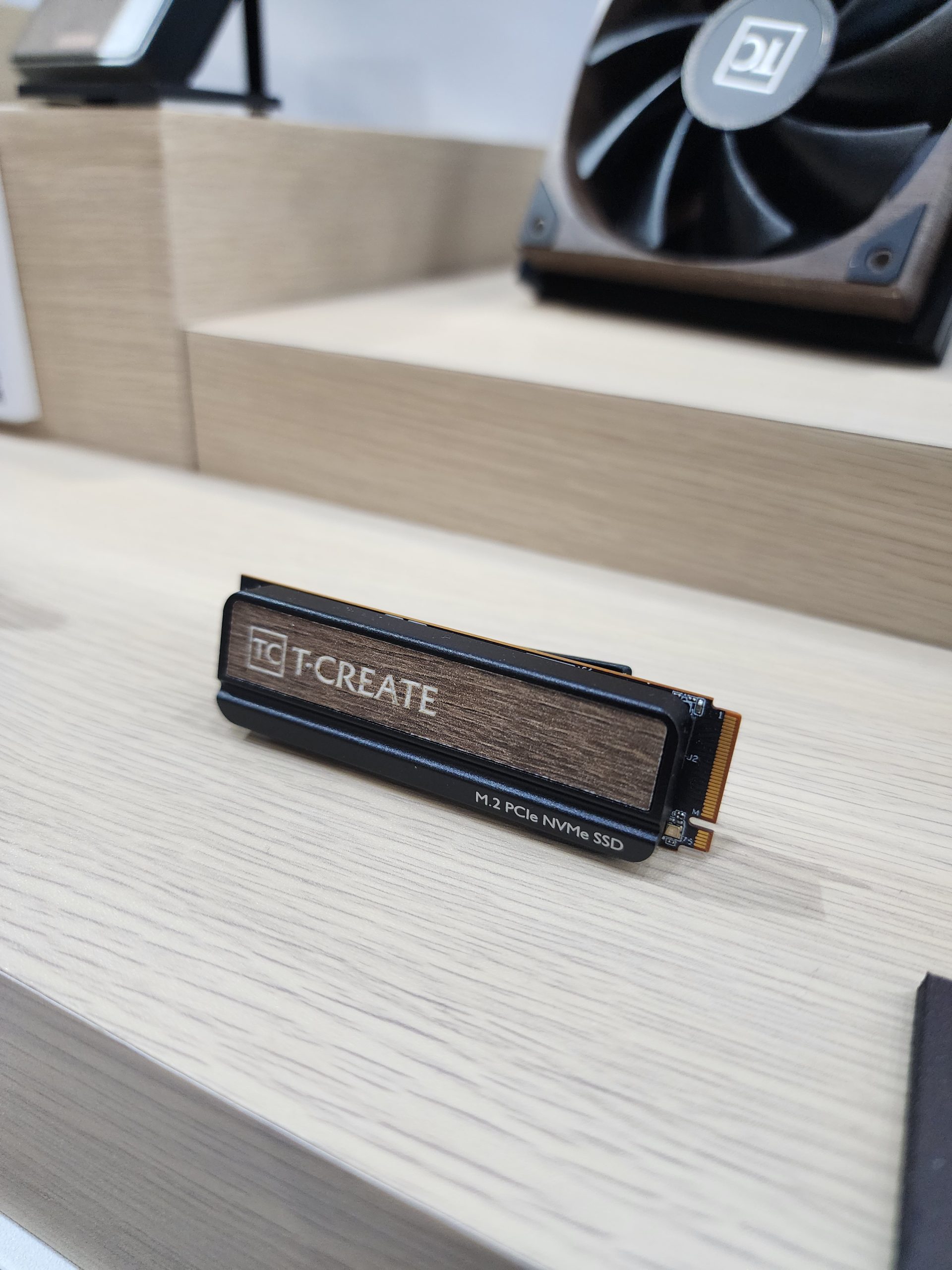 A TeamGroup T-Create M.2 PCIe NVMe SSD is displayed on a wooden surface with a T-Create-branded fan in the background.