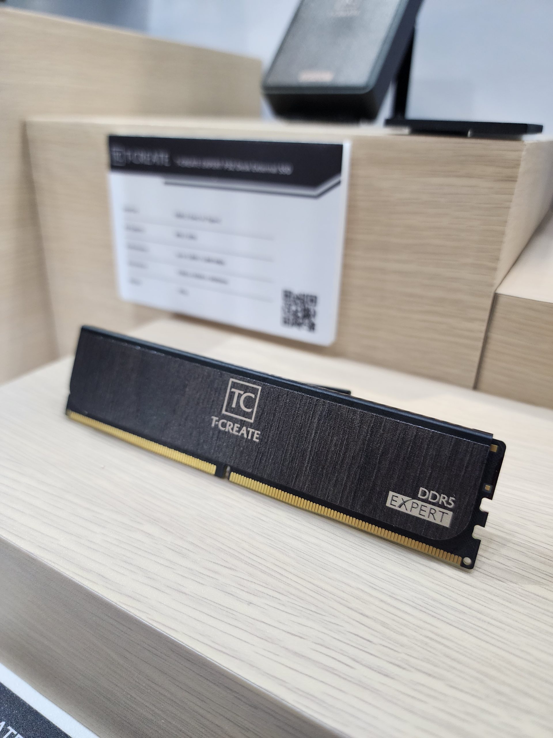 A T-CREATE DDR5 Expert RAM module is displayed on a wooden stand with product information in the background.