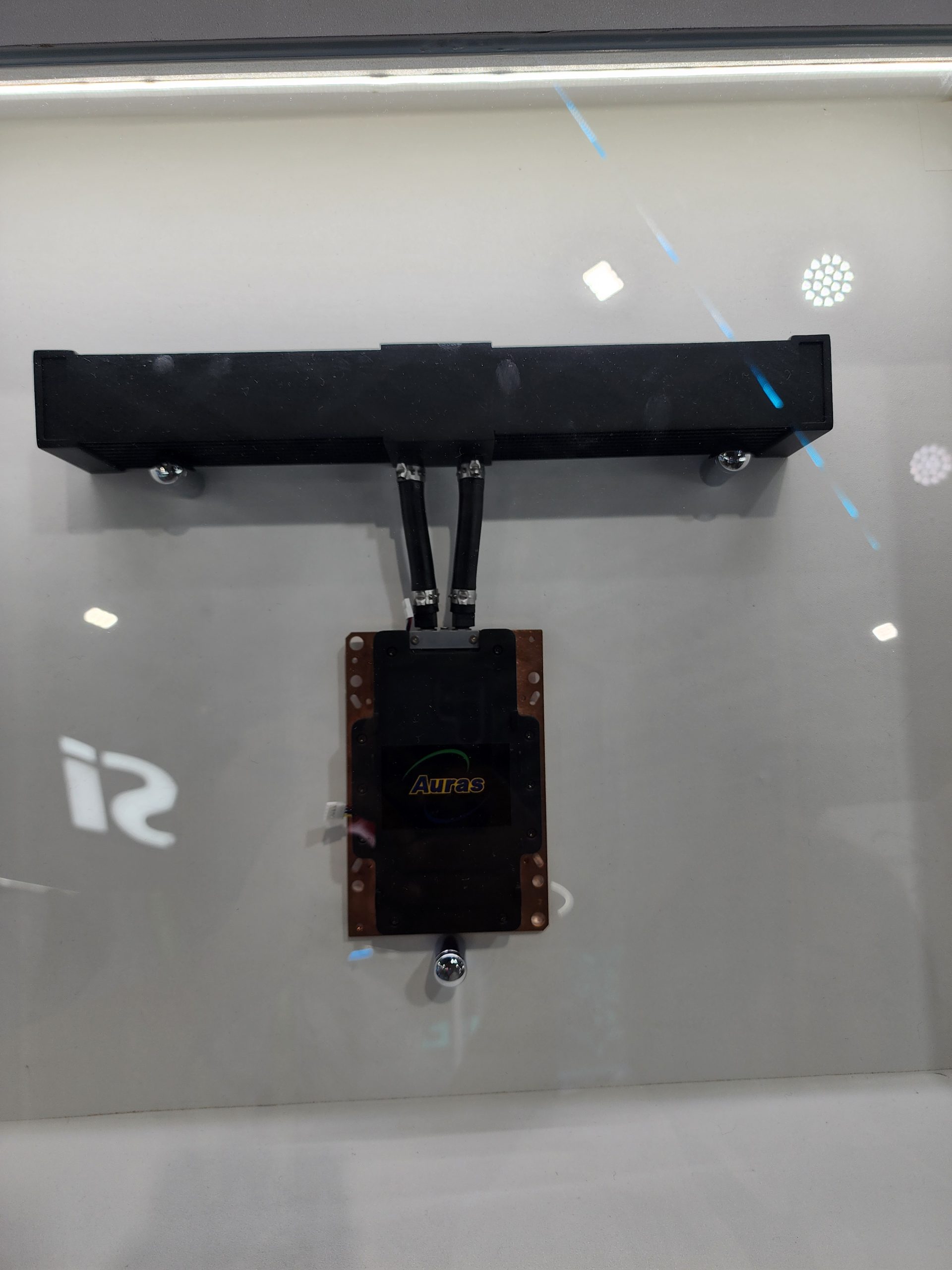 An Auras-branded liquid cooling component is displayed inside a glass case, featuring a black reservoir connected to a copper cold plate via two tubes.