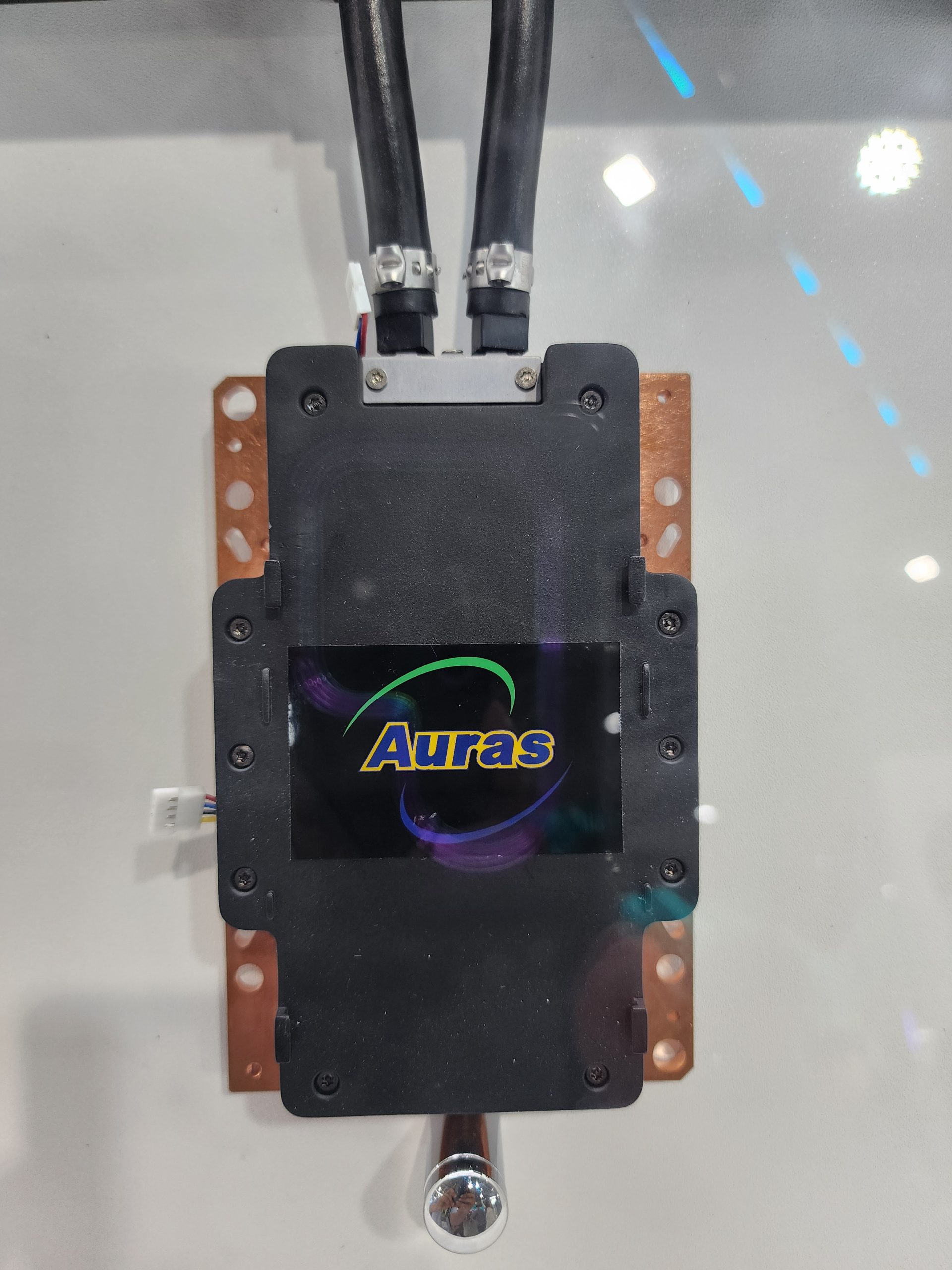 A liquid cooling block from Auras with two attached pipes is displayed on a metallic base.