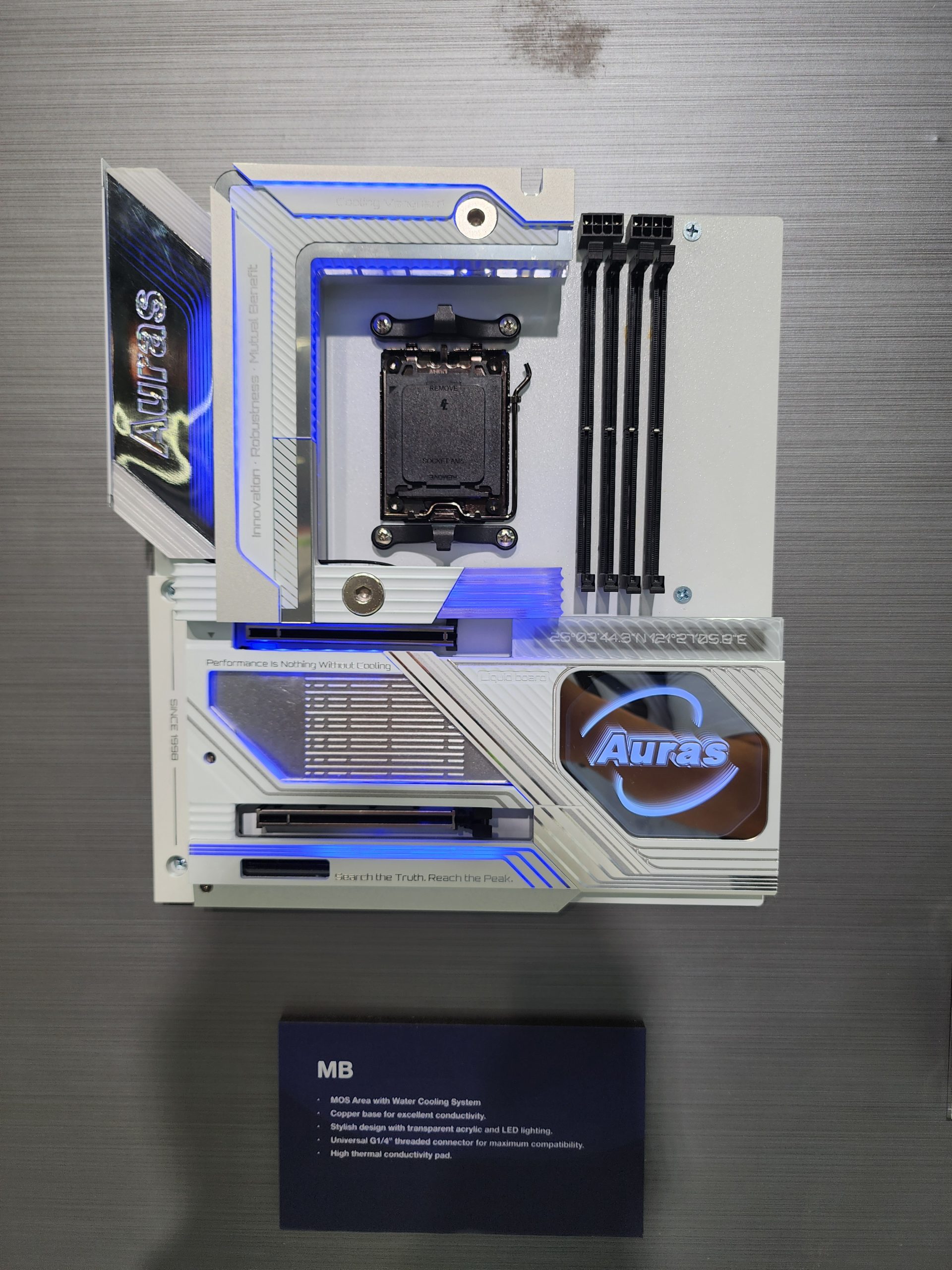 A white and blue Auras motherboard with LED lighting and the text 'Performance is Nothing Without Cooling' is displayed vertically with dimm slots and a CPU socket visible.