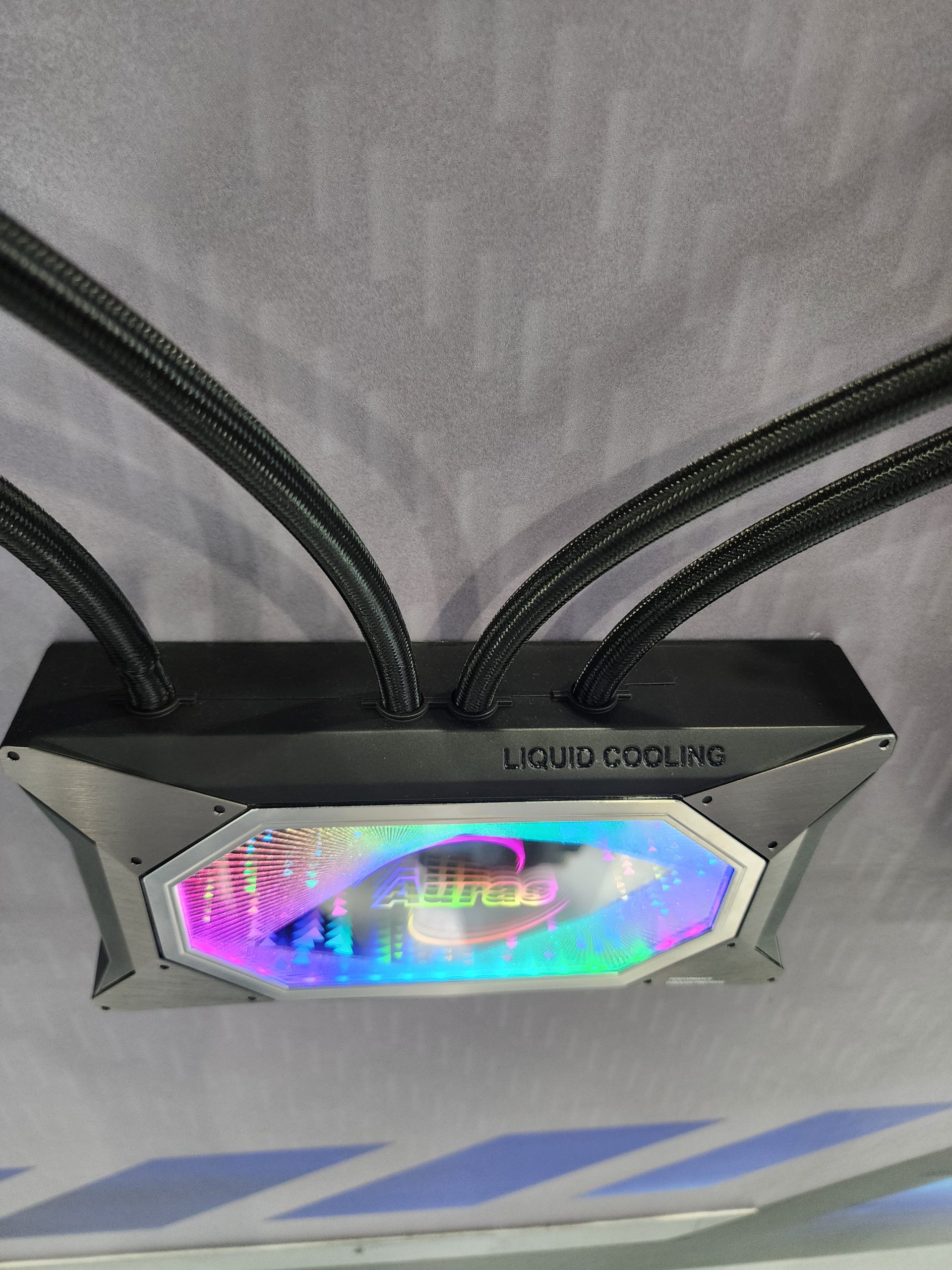 A CPU liquid cooling block with illuminated 'Aura' branding and the text 'LIQUID COOLING' on the side panel, featuring three connecting braided tubes.