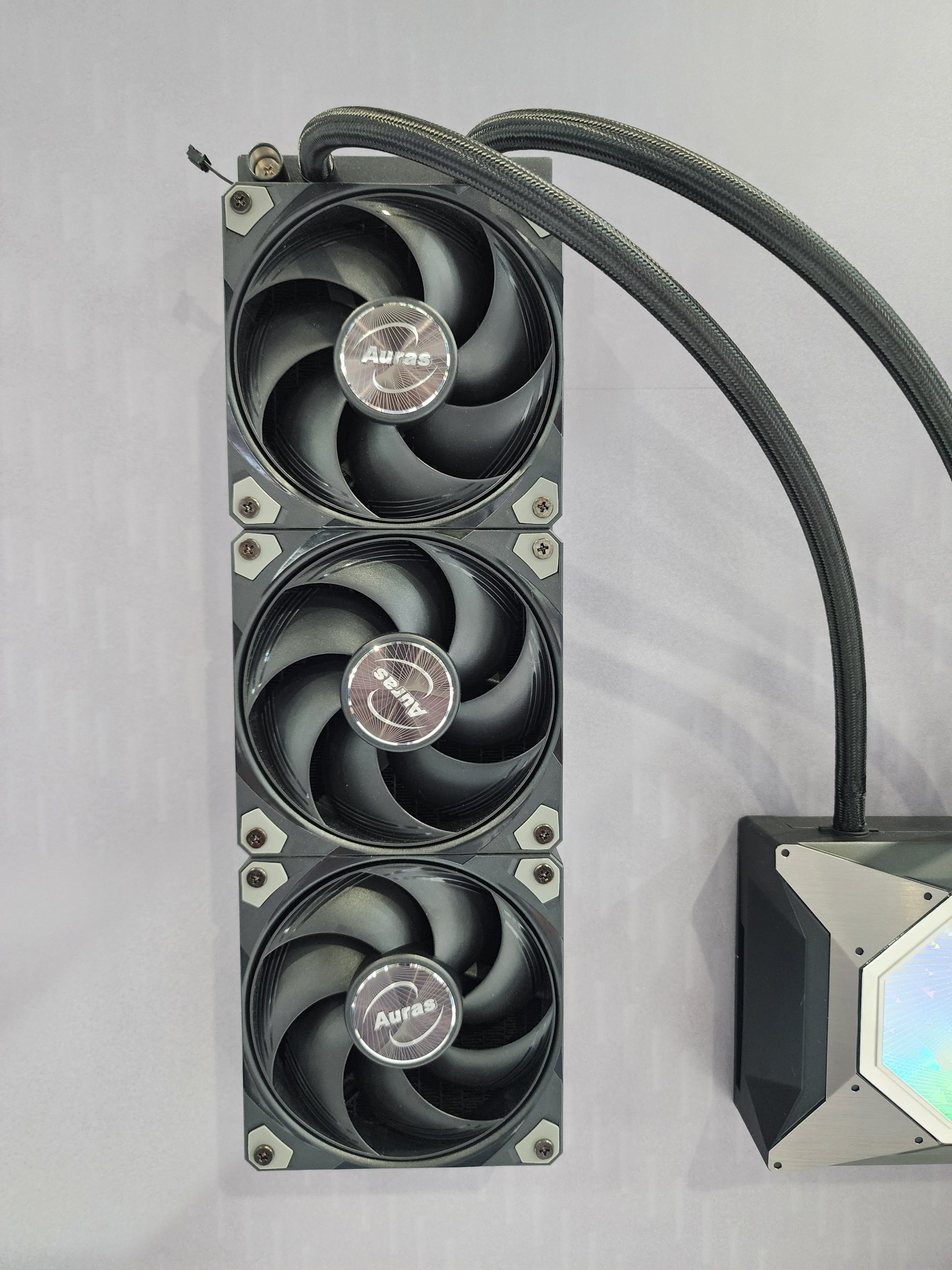 An Auras triple-fan cooling system with attached cables is mounted vertically on a surface.