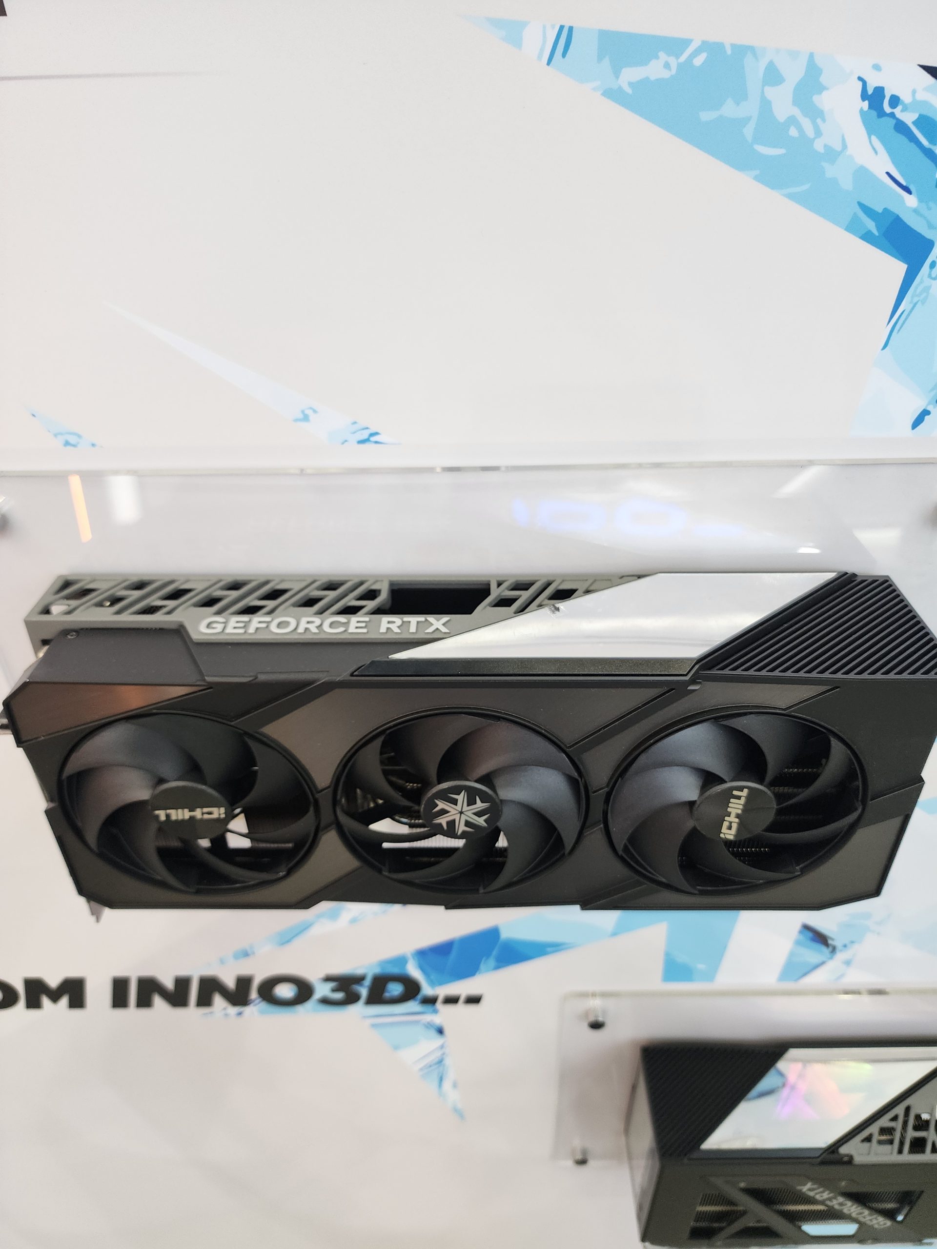 An Inno3D GeForce RTX graphics card with triple fans is displayed on a stand with a white and blue background.