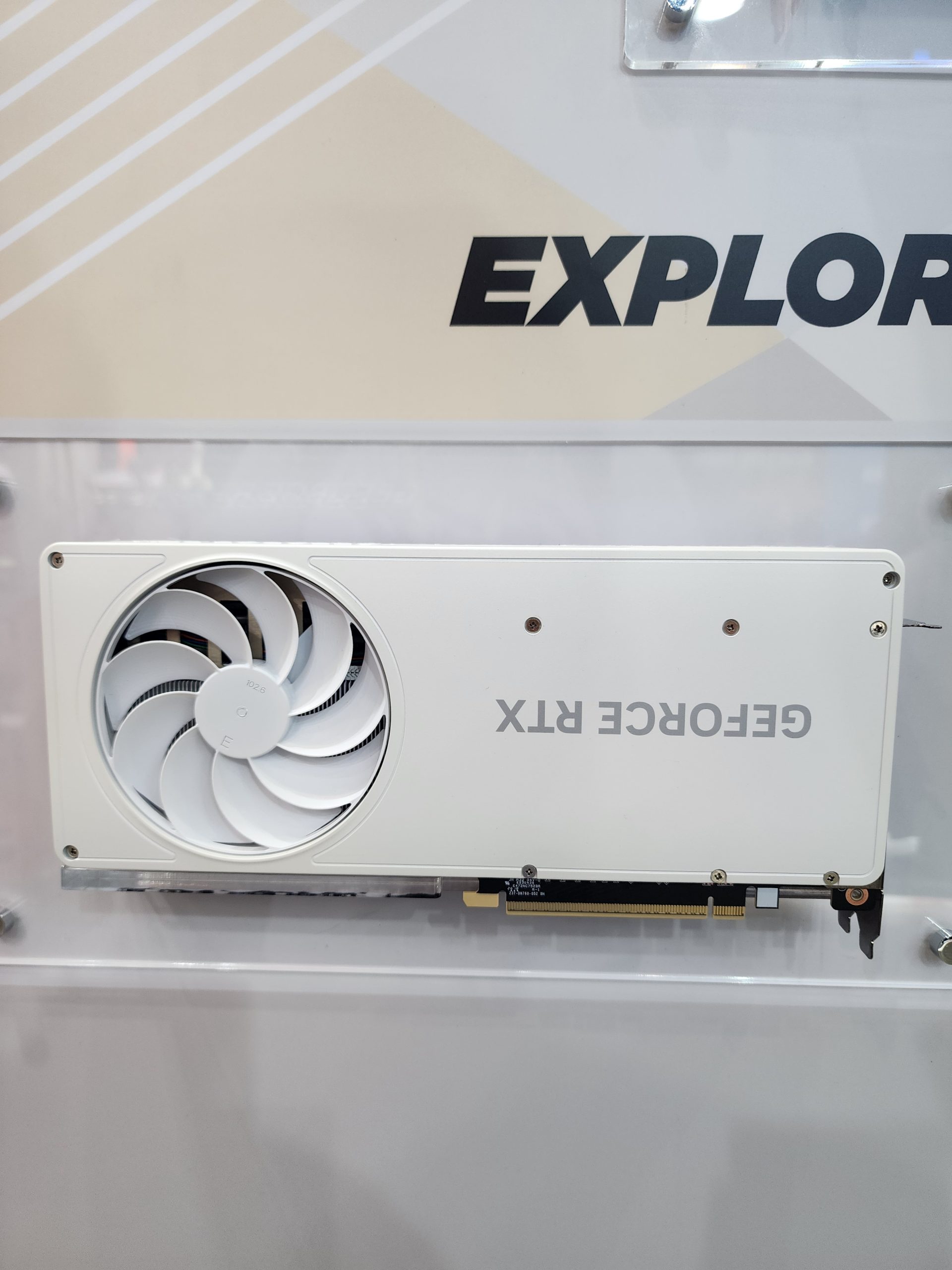 A white GeForce RTX graphics card is displayed vertically against a wall with 'EXPLORE' partially visible in the background.