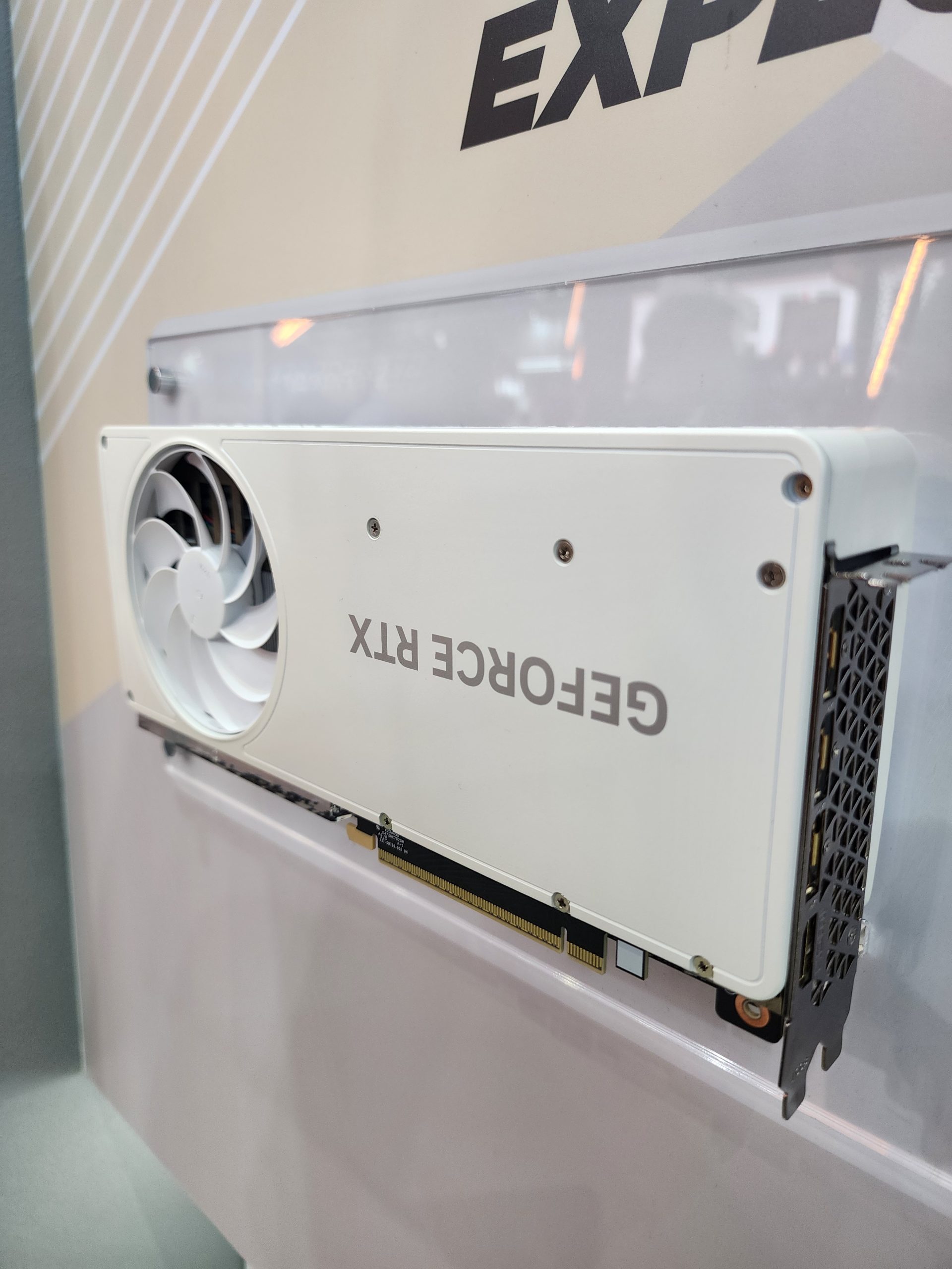 A white graphics card labeled GeForce RTX with a single fan displayed against a wall with part of the word EXPERIENCE visible.
