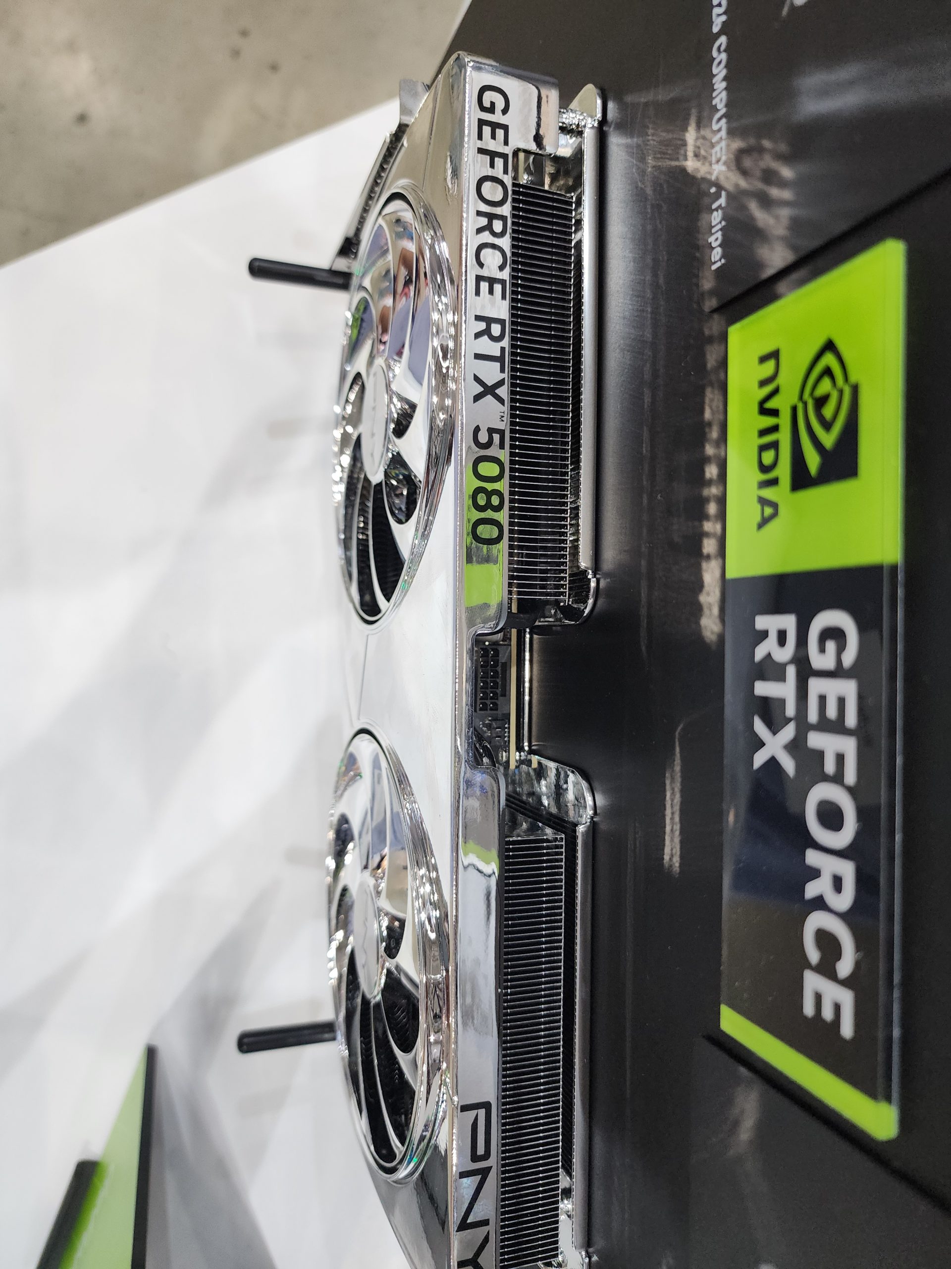 A PNY NVIDIA GeForce RTX 5080 graphics card with dual fans is displayed vertically next to NVIDIA-branded packaging.