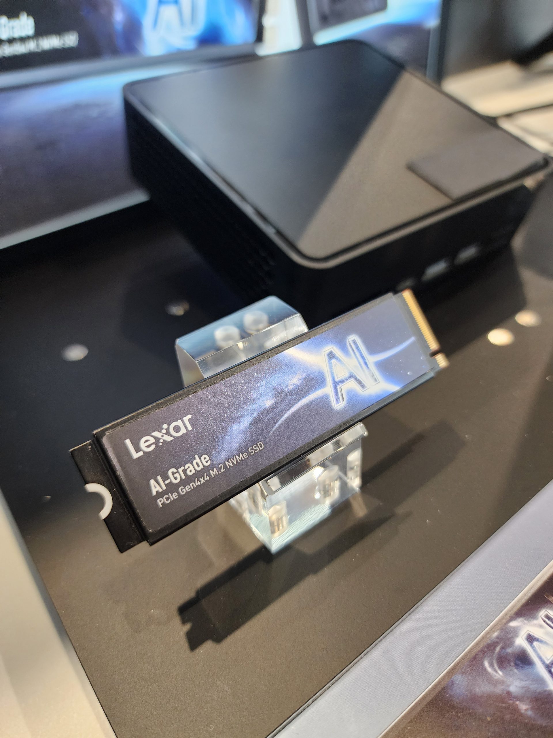 A Lexar PCIe Gen4x4 M.2 NVMe SSD labeled 'AI-Grade' is displayed on a stand next to a black compact PC case.