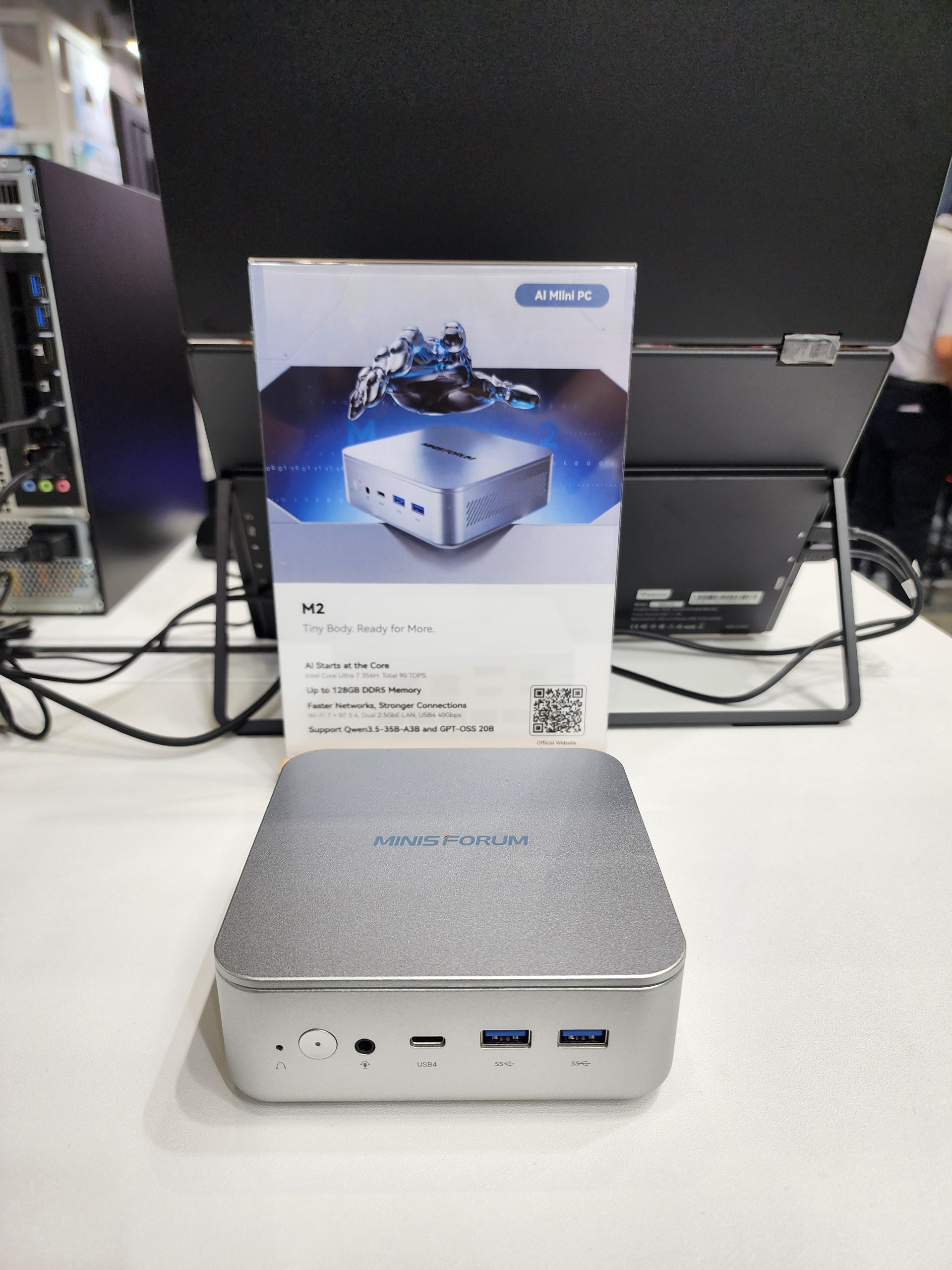 A Minisforum M2 AI Mini PC is displayed on a table next to promotional material highlighting features like 'AI Starts at the Core' and 'Up to 128GB DDR5 Memory'.