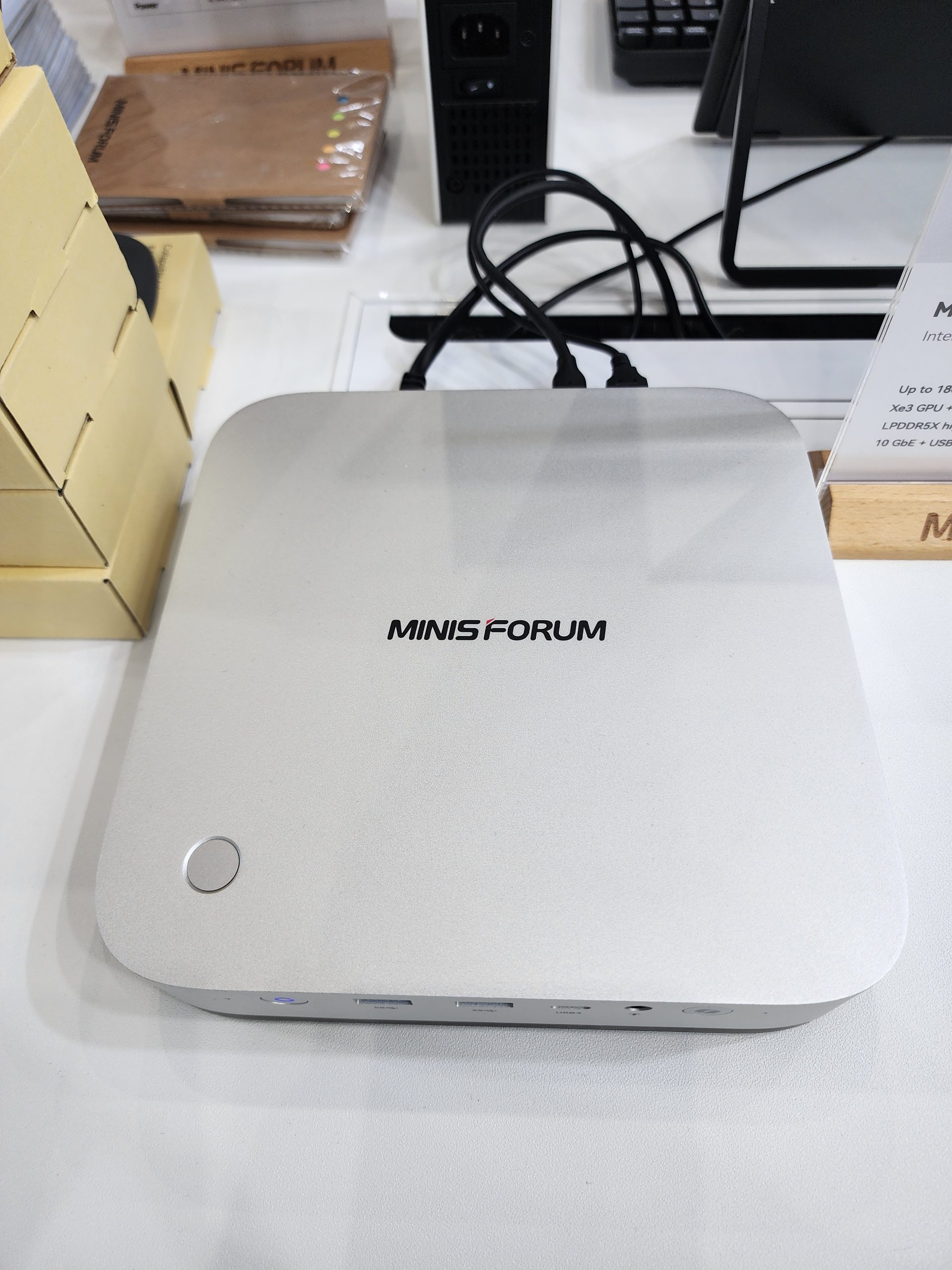 A silver Minisforum mini PC is displayed on a white table, featuring front ports and a power button.