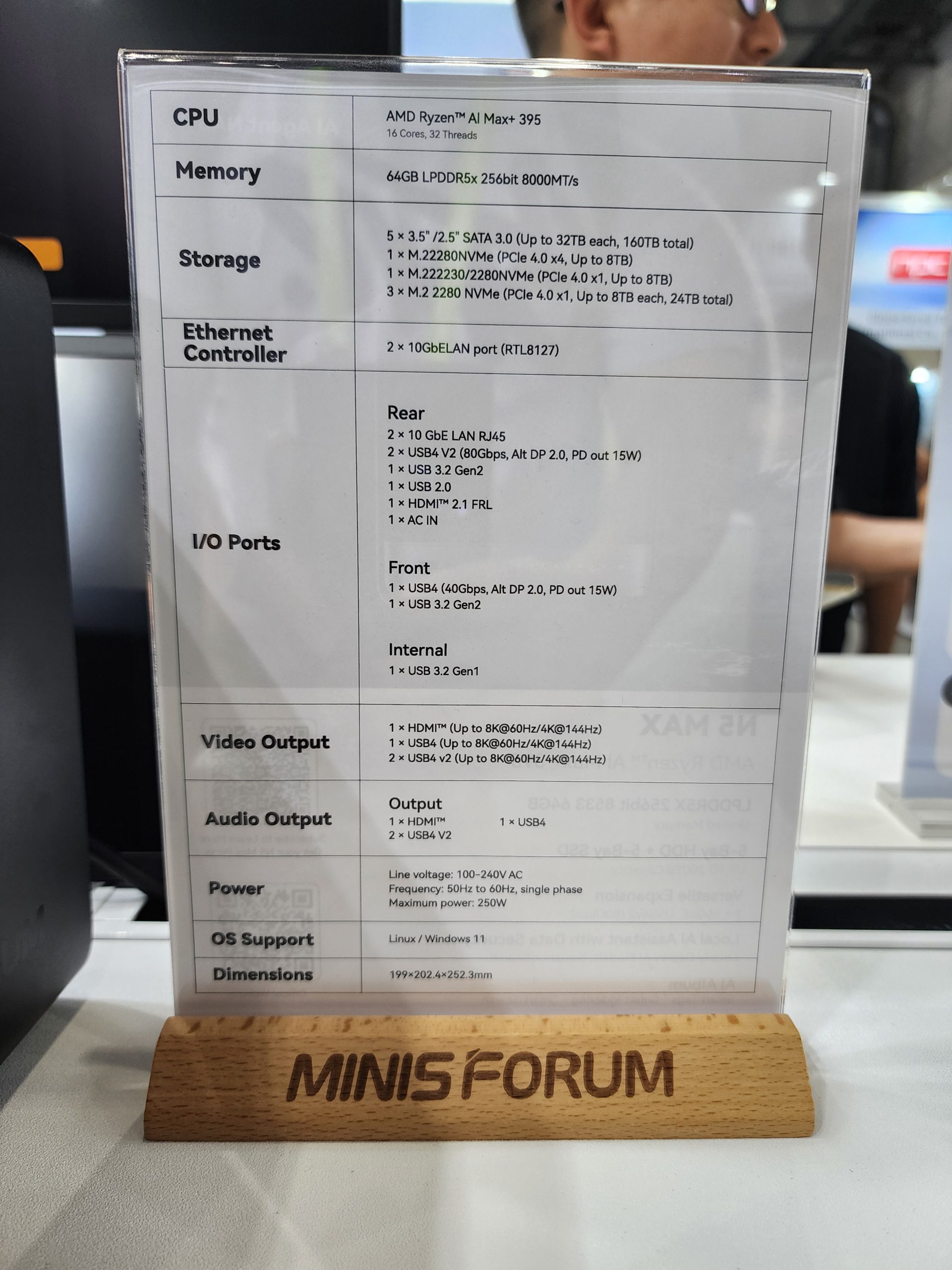 A specifications sheet on a wooden stand labeled Minisforum details the features of an AMD Ryzen AI Max 395 with 64GB LPDDR5x, various storage options including M.2 NVMe, and 10GbE LAN ports.