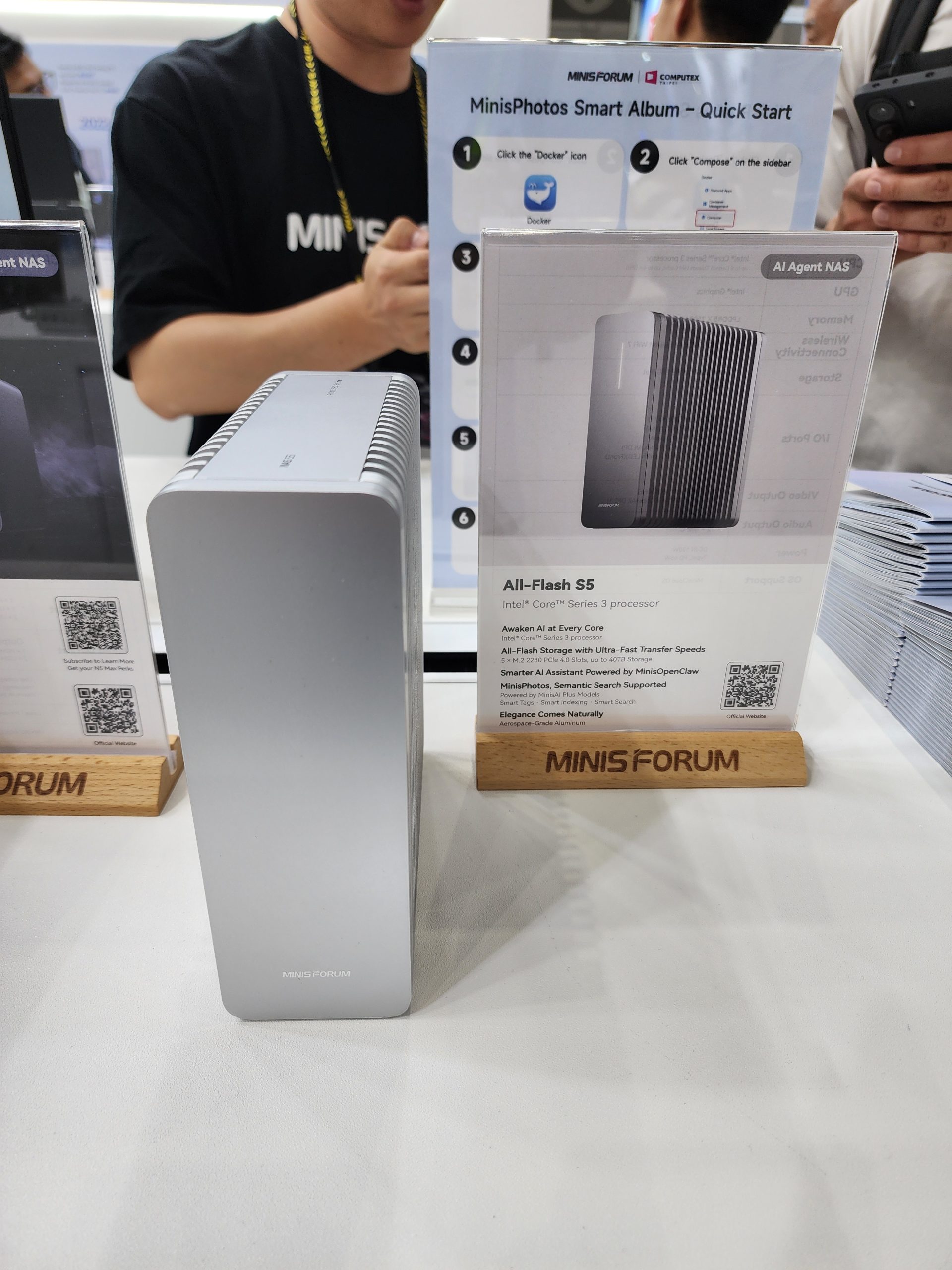 A MinisForum All-Flash S5 device is displayed on a table with a sign detailing its Intel Core Series 3 processor capabilities, while a board titled 'MinisPhotos Smart Album - Quick Start' is in the background.