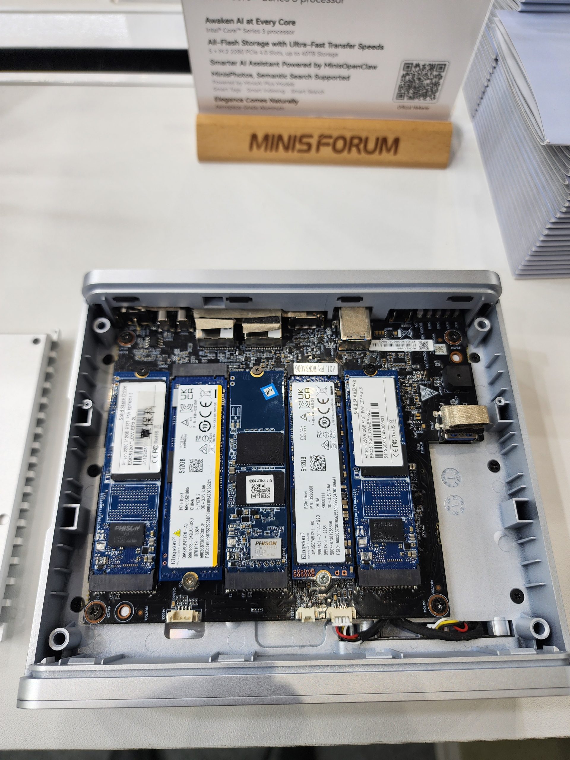 The Minisforum case is open to reveal three NVMe storage drives installed on the motherboard, with a sign in the background reading 'MINISFORUM'.