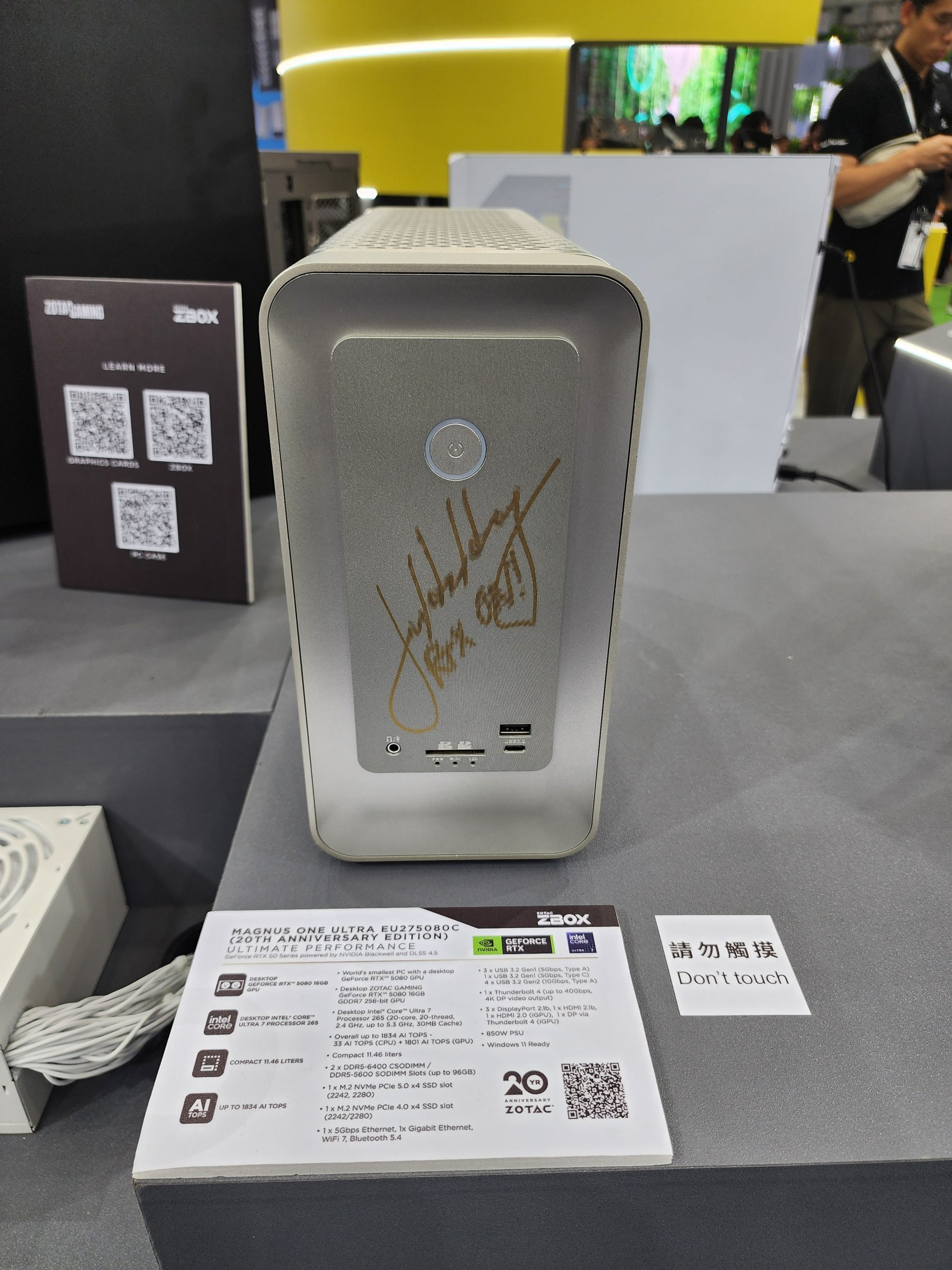 ZOTAC Magnus One Ultra EU27580C, a 20th Anniversary Edition desktop, is displayed with a signature on the front panel and a specification sheet highlighting Intel Core 13th Gen and NVIDIA GeForce RTX 4070, alongside a 'Don't touch' sign.