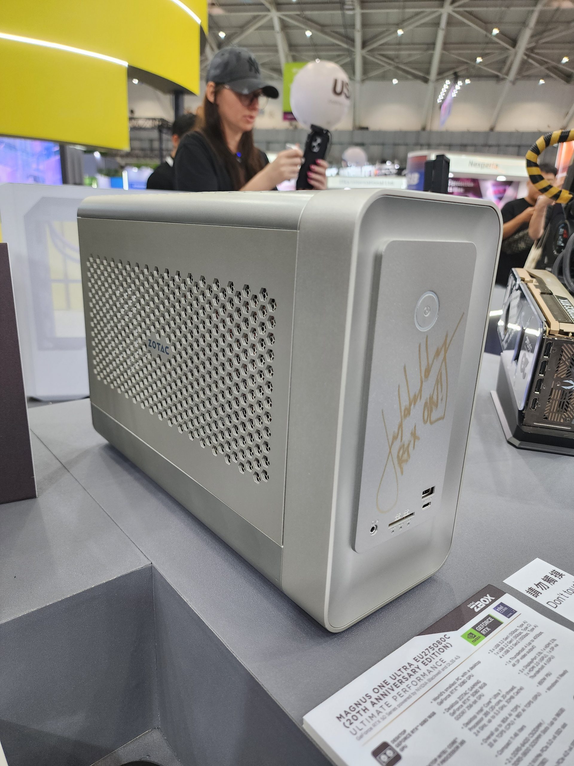 A ZOTAC Magnus One Ultra EU1Z590C mini PC is displayed at an event, featuring a '20th Anniversary Edition' label and a signature on the front panel.