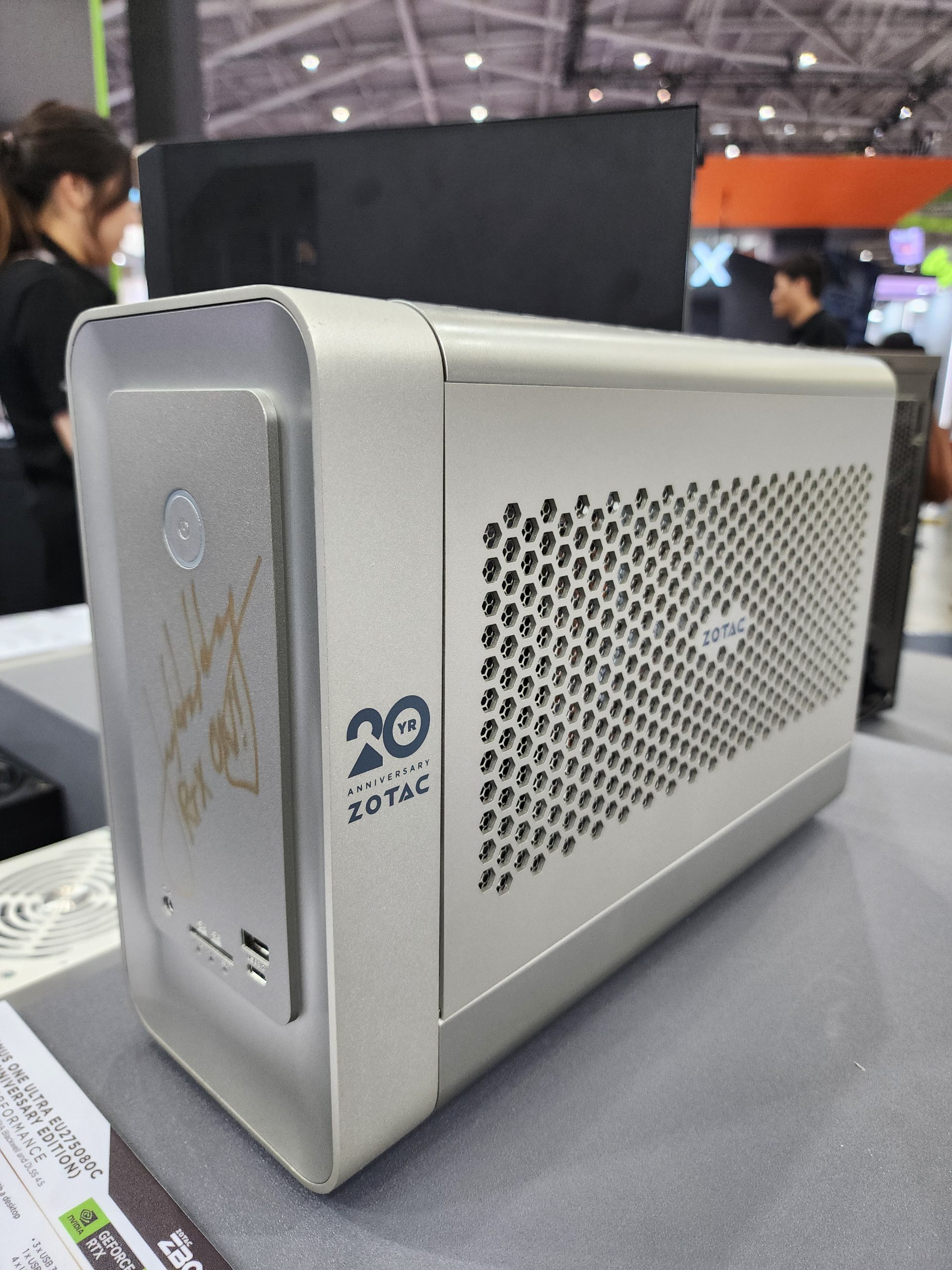The image shows a Zotac 20th Anniversary Edition mini-PC case on display, featuring a silver design with ventilation holes and USB ports at the front.