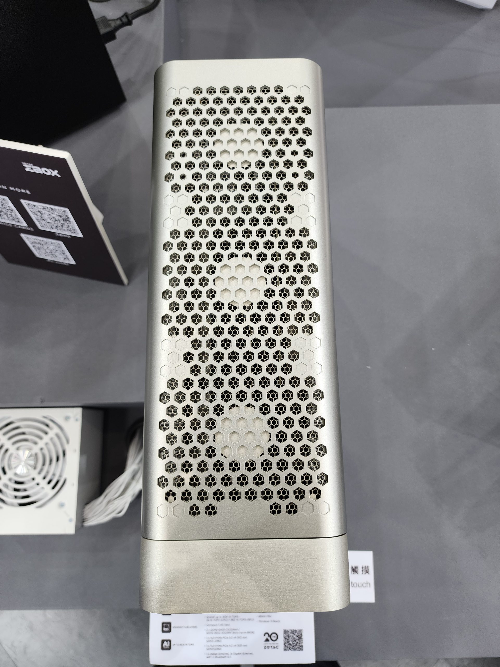 A ZOTAC ZBOX mini PC with a hexagonal perforated design on a table, accompanied by a separate cooling fan unit.
