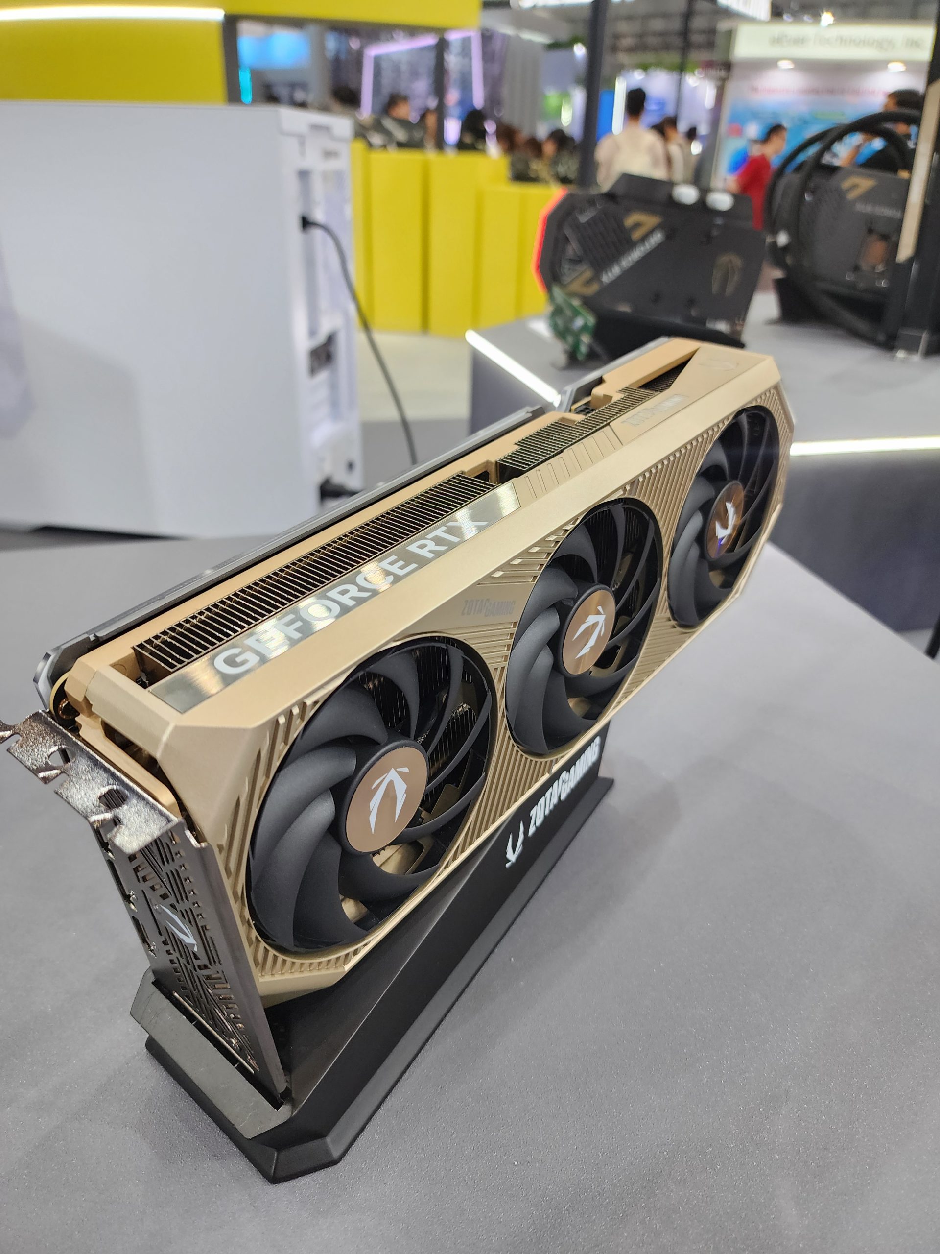 A Zotac Gaming GeForce RTX graphics card with a gold-colored design is displayed on a stand, showing three cooling fans in a busy exhibition hall.