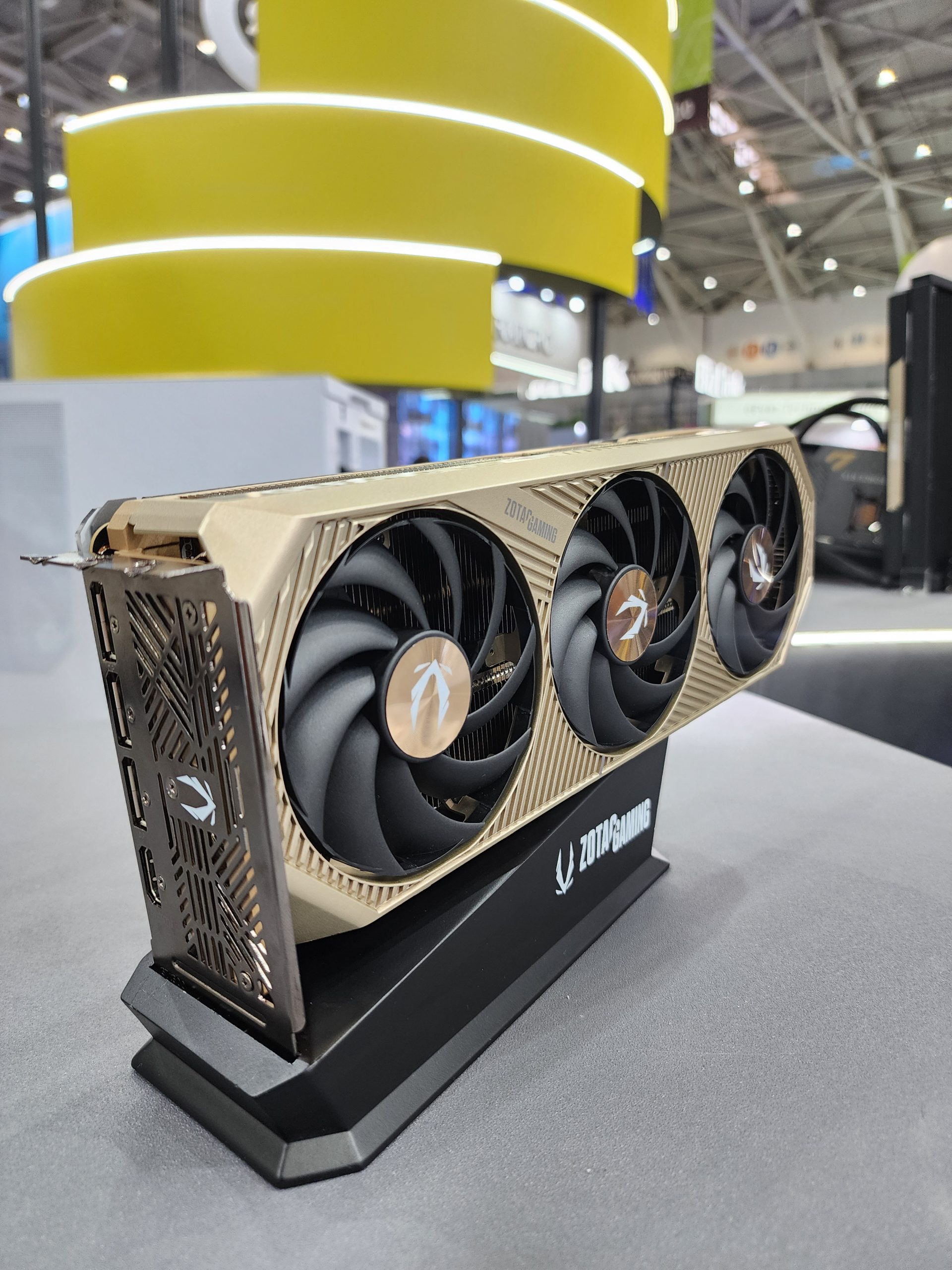 A ZOTAC GAMING graphics card with triple fans and a gold-colored design is displayed on a stand in an exhibition setting.