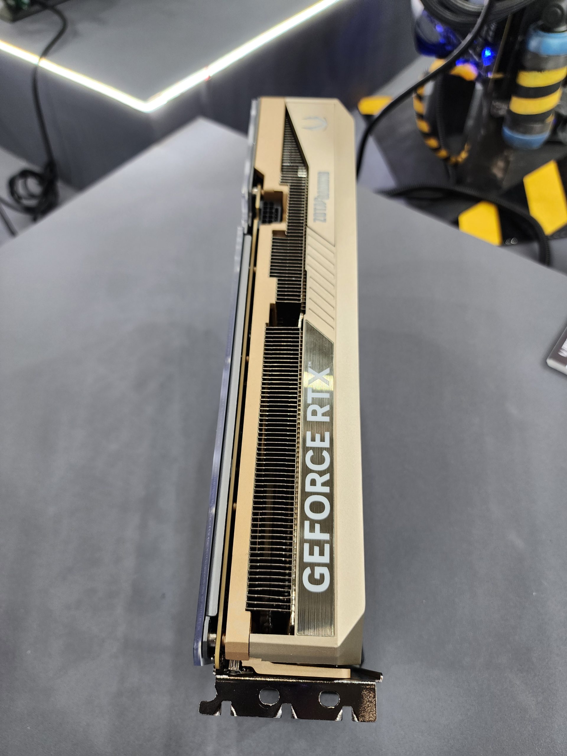 A Zotac GeForce RTX graphics card is displayed in a beige finish with a visible fan and heat sinks on a tabletop.