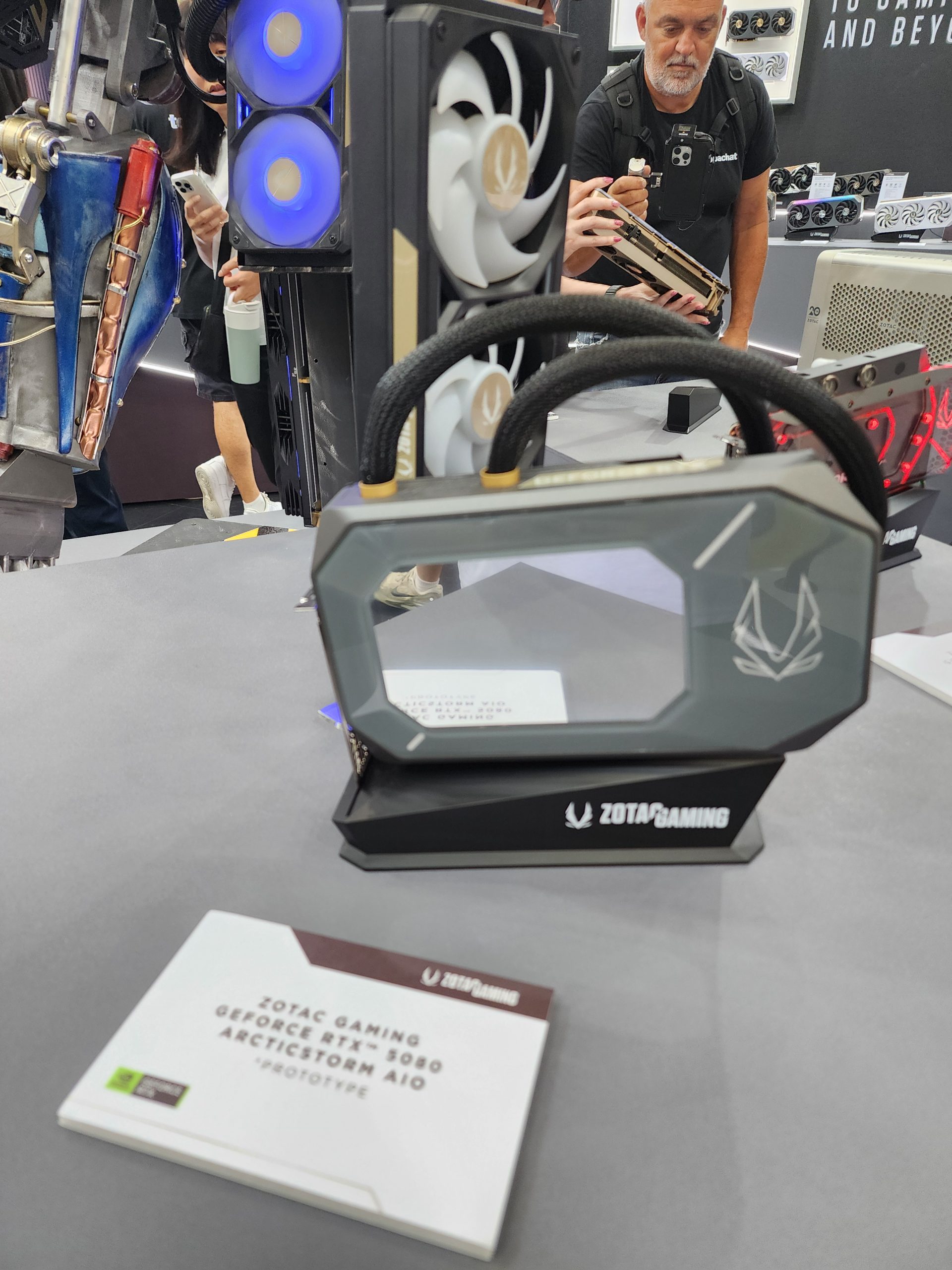 A Zotac Gaming GeForce RTX 5090 ArcticStorm AIO prototype is displayed on a table with a label in the foreground and computer cooling components in the background.