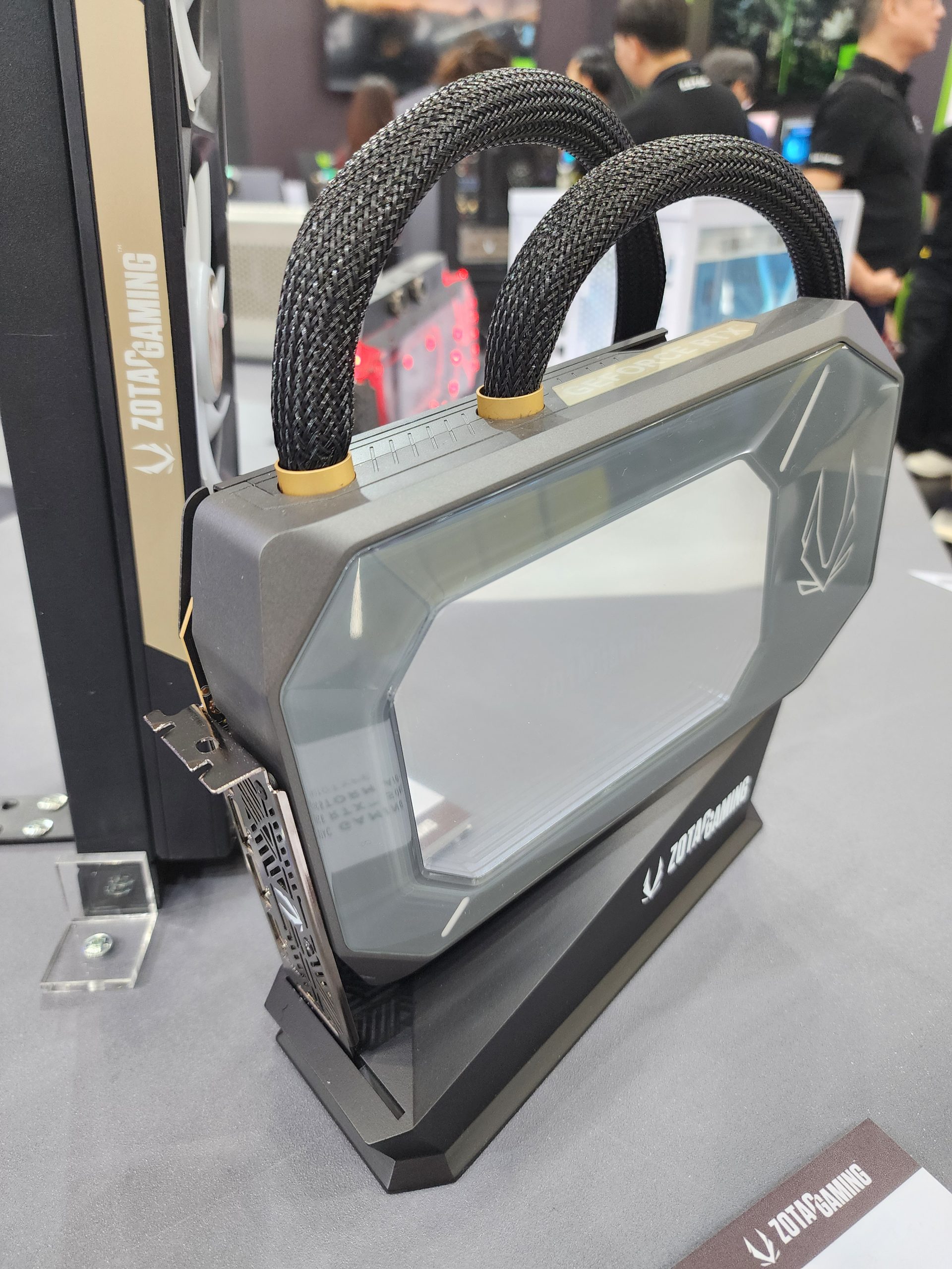 A ZOTAC GAMING graphics card with braided power cables is displayed vertically on a stand at an expo booth.