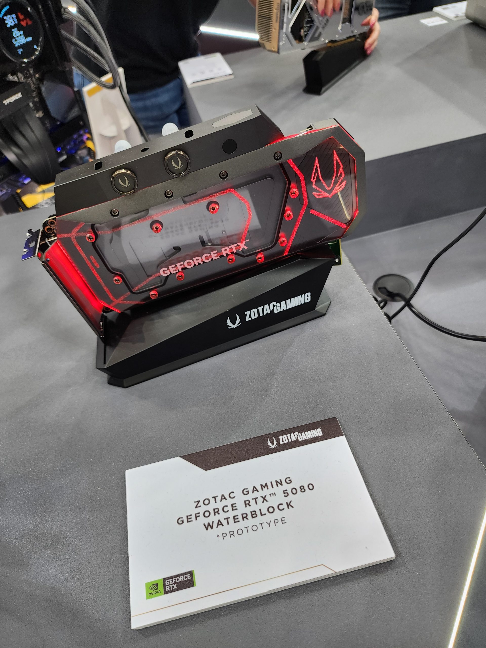 A Zotac Gaming GeForce RTX 5080 graphics card prototype with a waterblock is displayed, featuring red LED lighting and the Zotac logo.