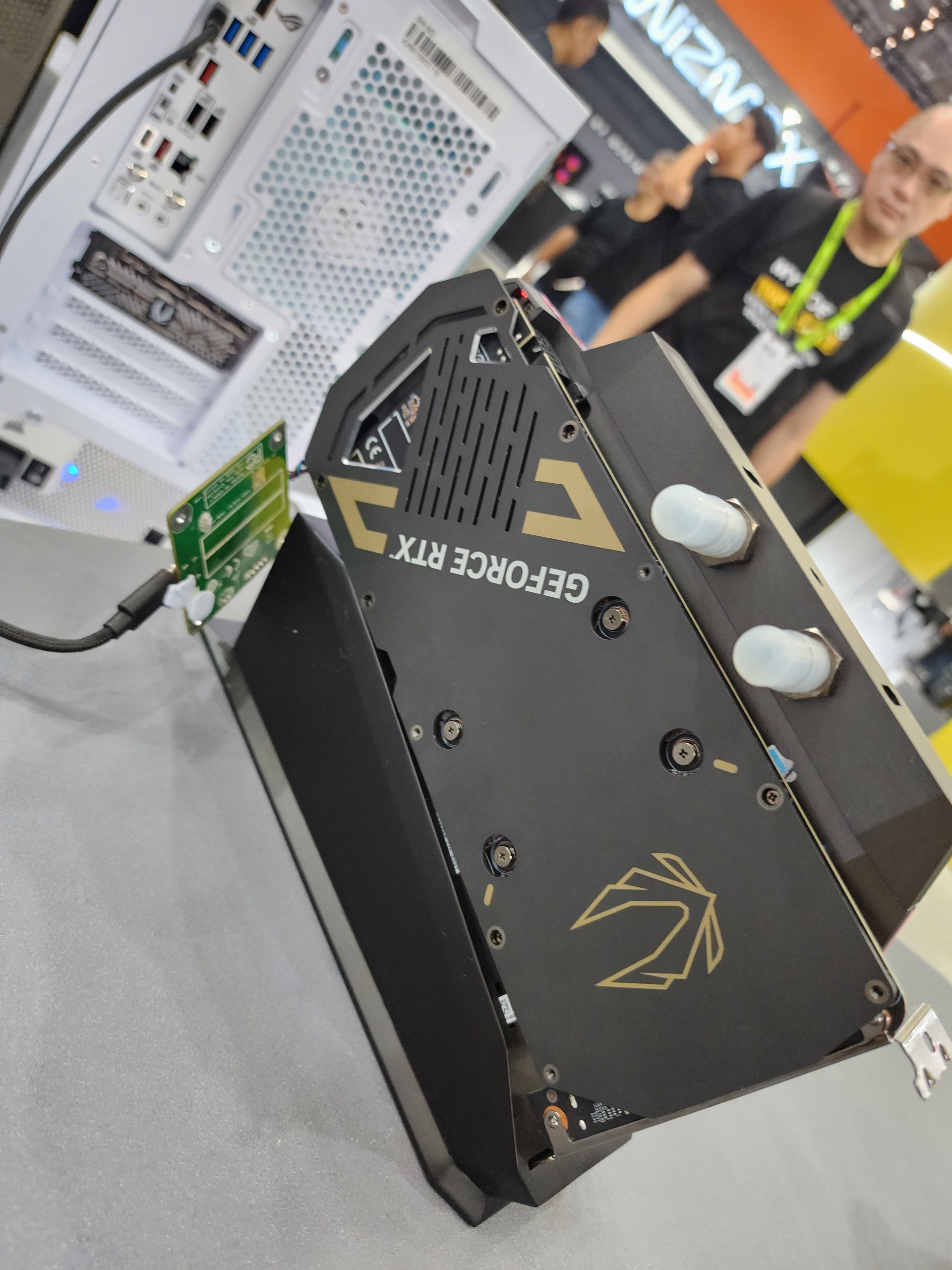 A Zotac graphics card with visible 'GEFORCE RTX' branding is mounted vertically next to a white PC case with multiple rear ports, observed in a tech expo setting with attendees in the background.