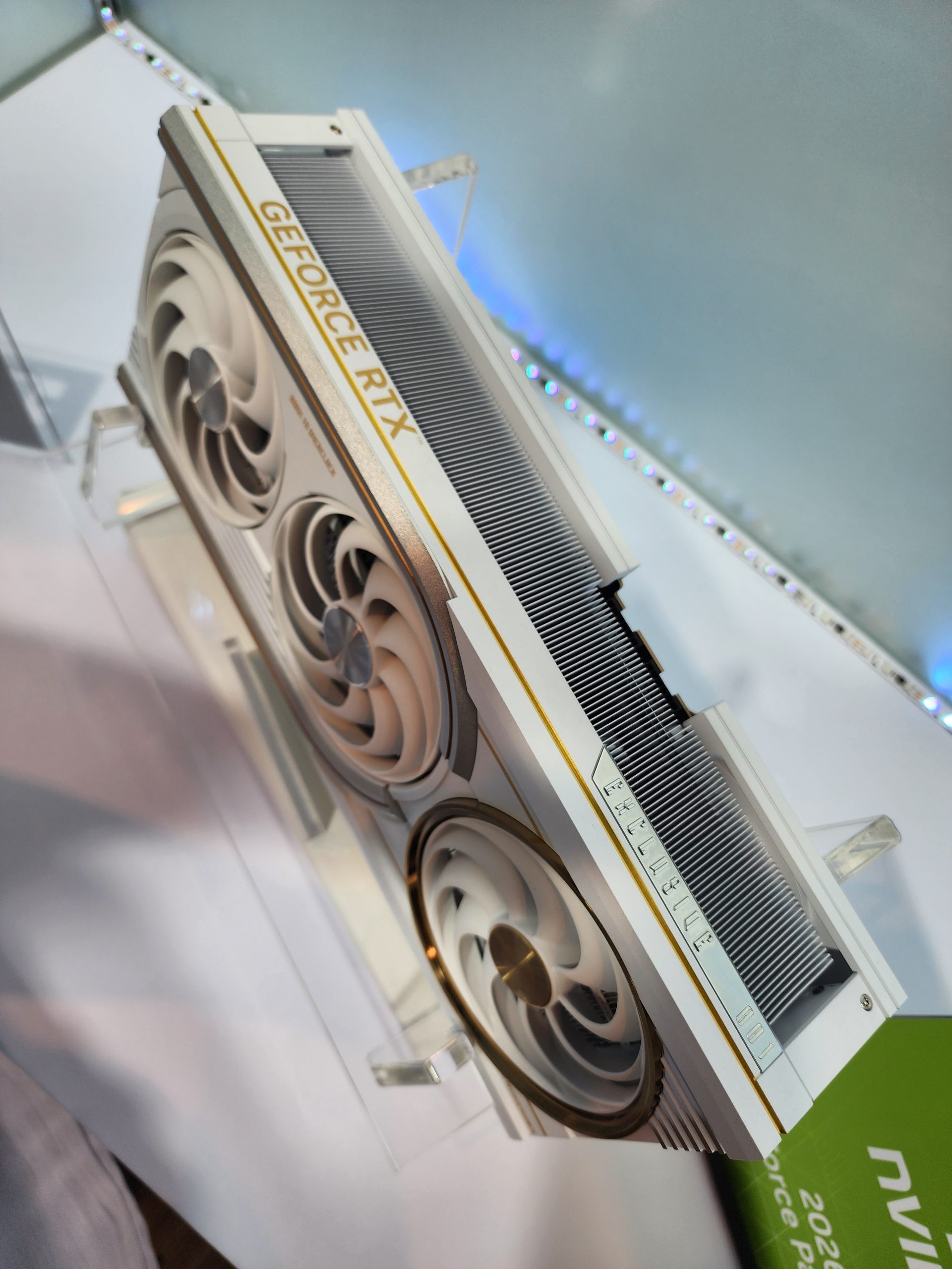 A white NVIDIA GeForce RTX graphics card with triple fans is displayed on a clear stand.