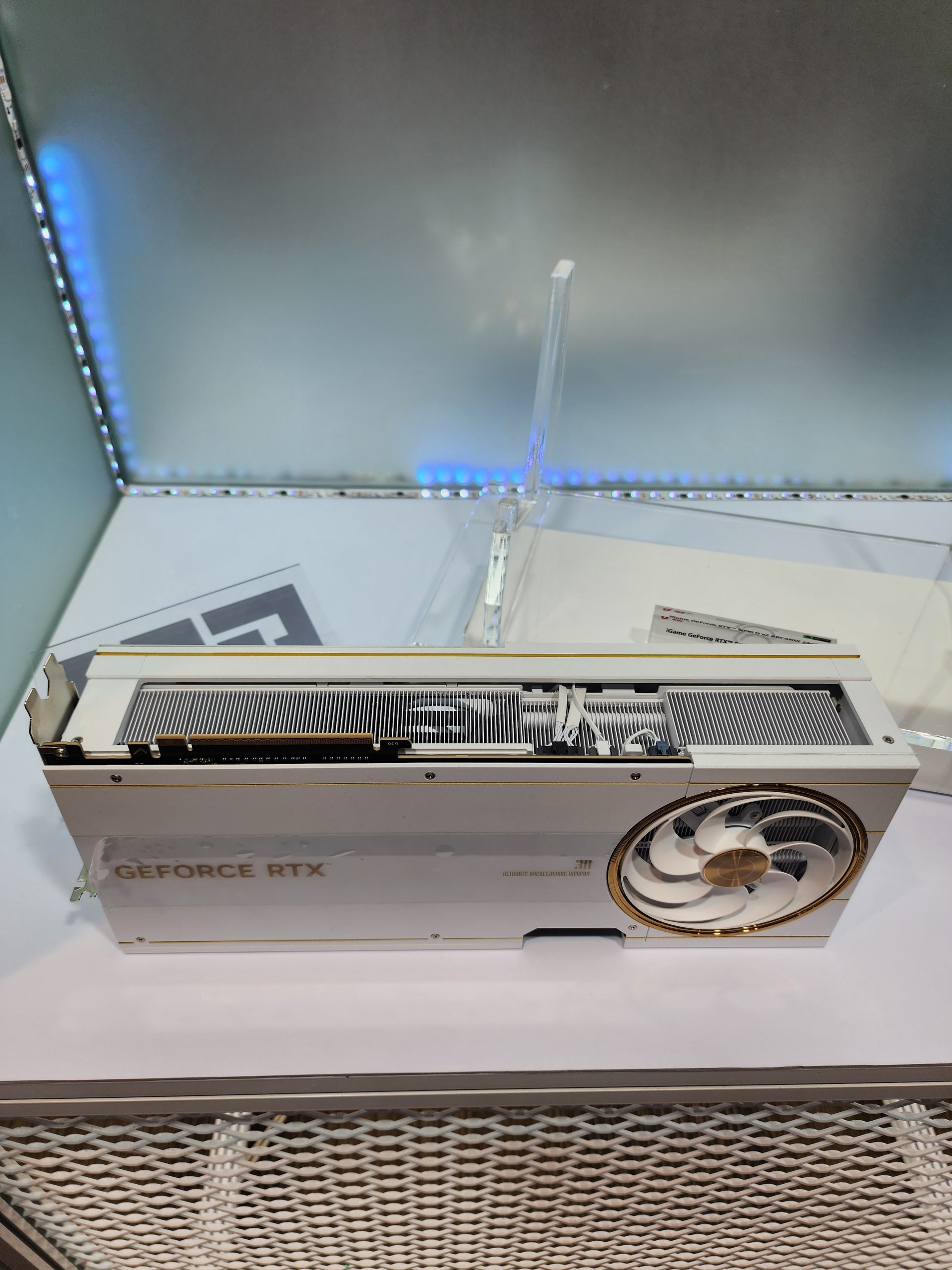 A white and gold NVIDIA GeForce RTX graphics card is displayed on a stand, featuring a large cooling fan and visible 'GEFORCE RTX' branding.