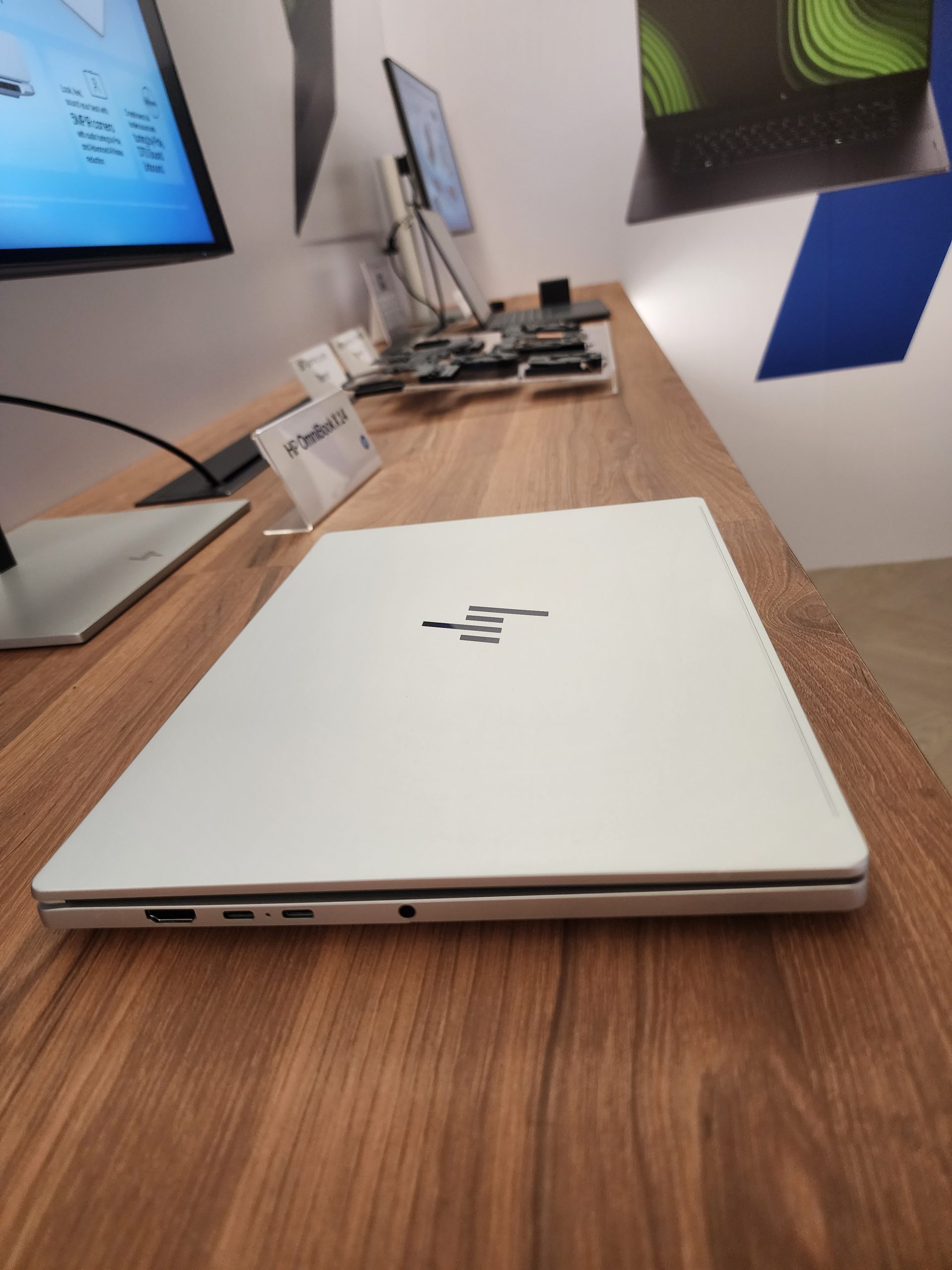 An HP EliteBook x360 is displayed partially closed on a wooden table next to a sign labeled 'HP EliteBook 1020,' surrounded by monitors and electronic components.