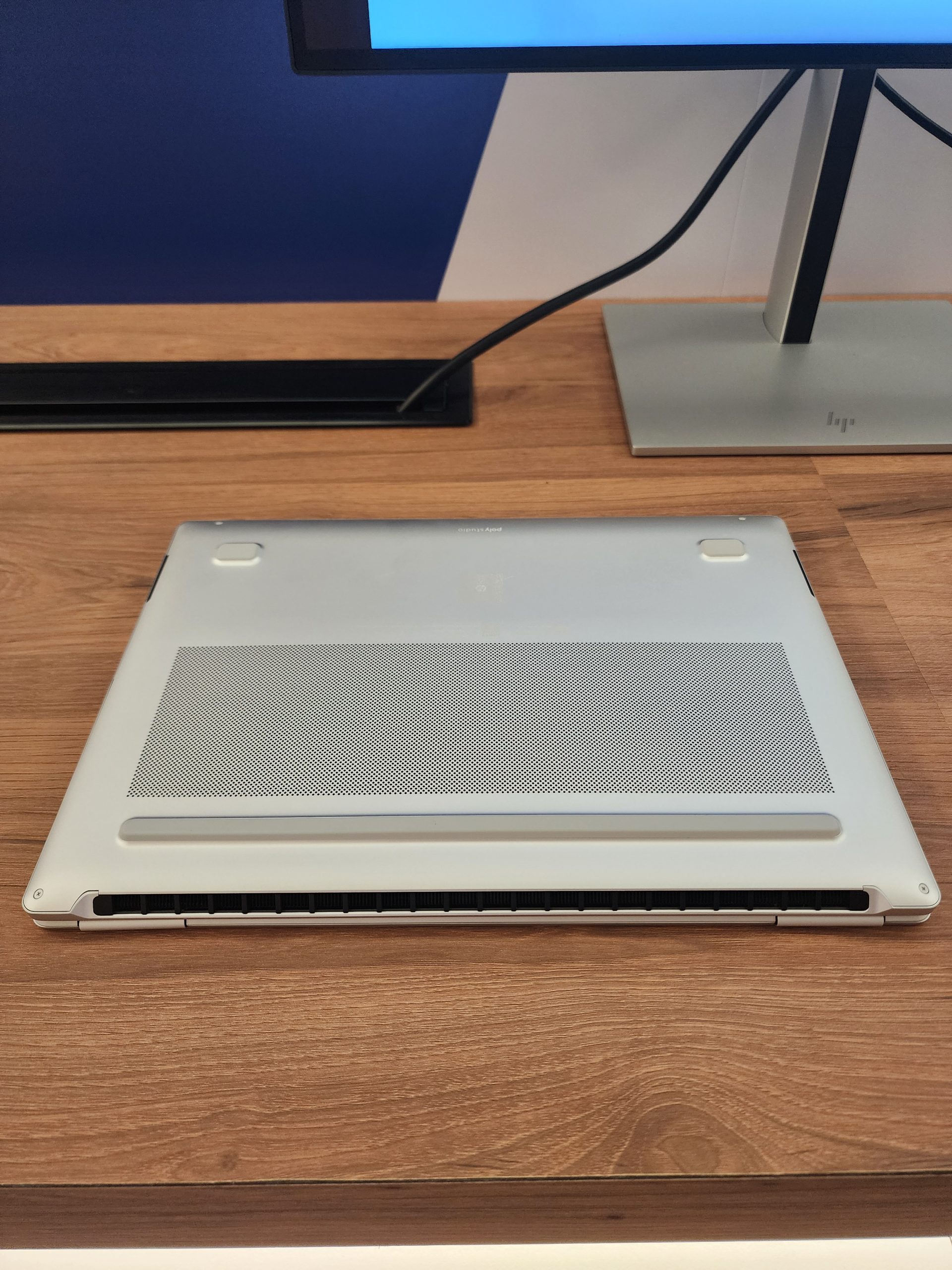 The underside of a silver unbranded laptop on a wooden desk, with a nearby HP monitor stand visible.