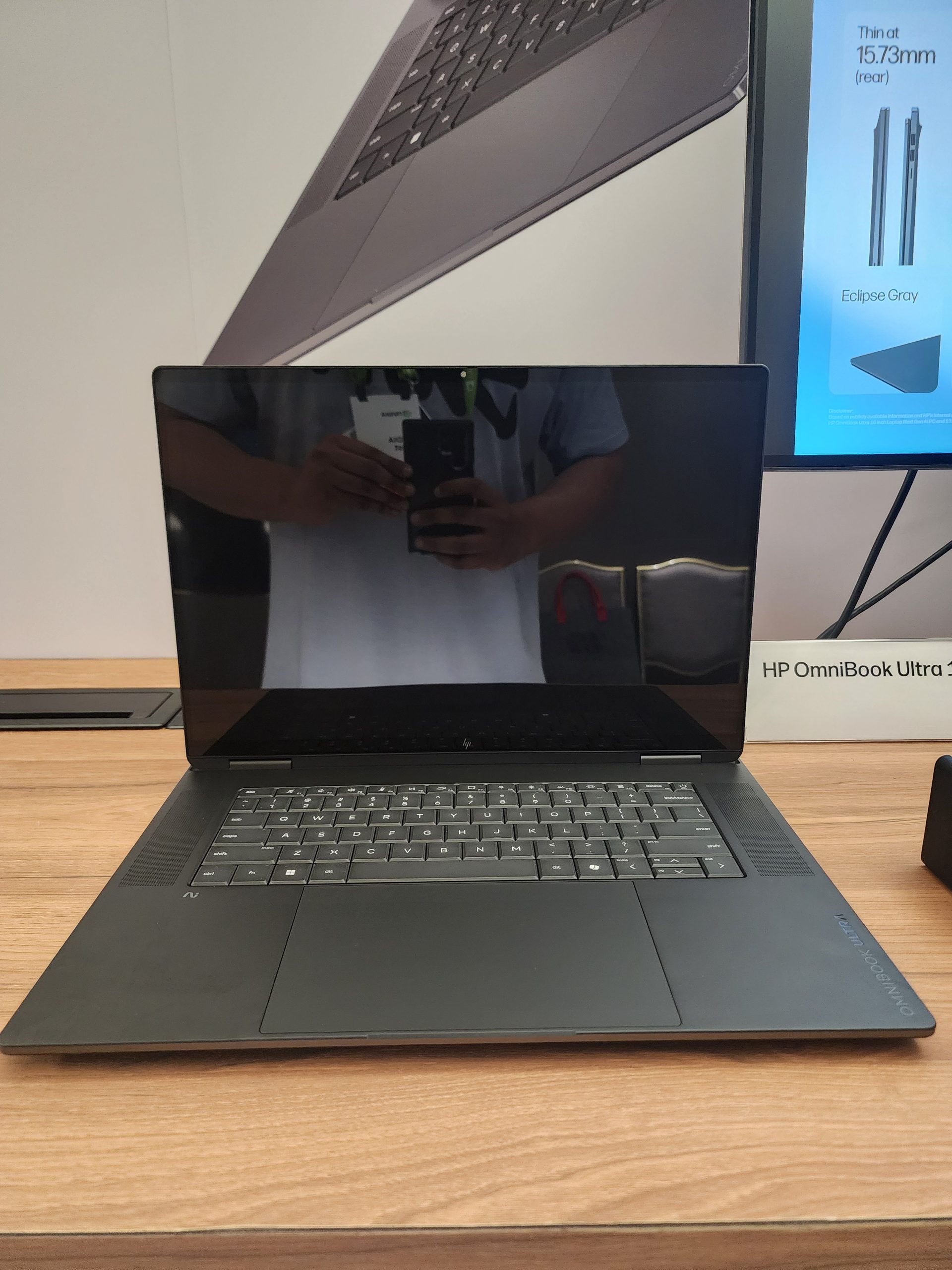 The HP OmniBook Ultra V laptop is displayed on a wooden table, with a promotional poster in the background showing the device's thin profile labeled as '15.73mm' in 'Eclipse Gray' color.