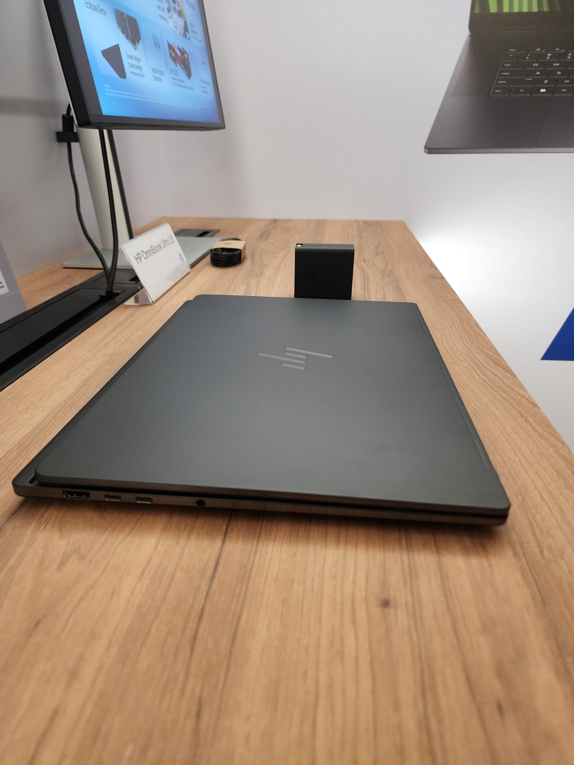 An HP Dragonfly G4 laptop is closed on a wooden desk, with a monitor displaying tech product slides in the background.
