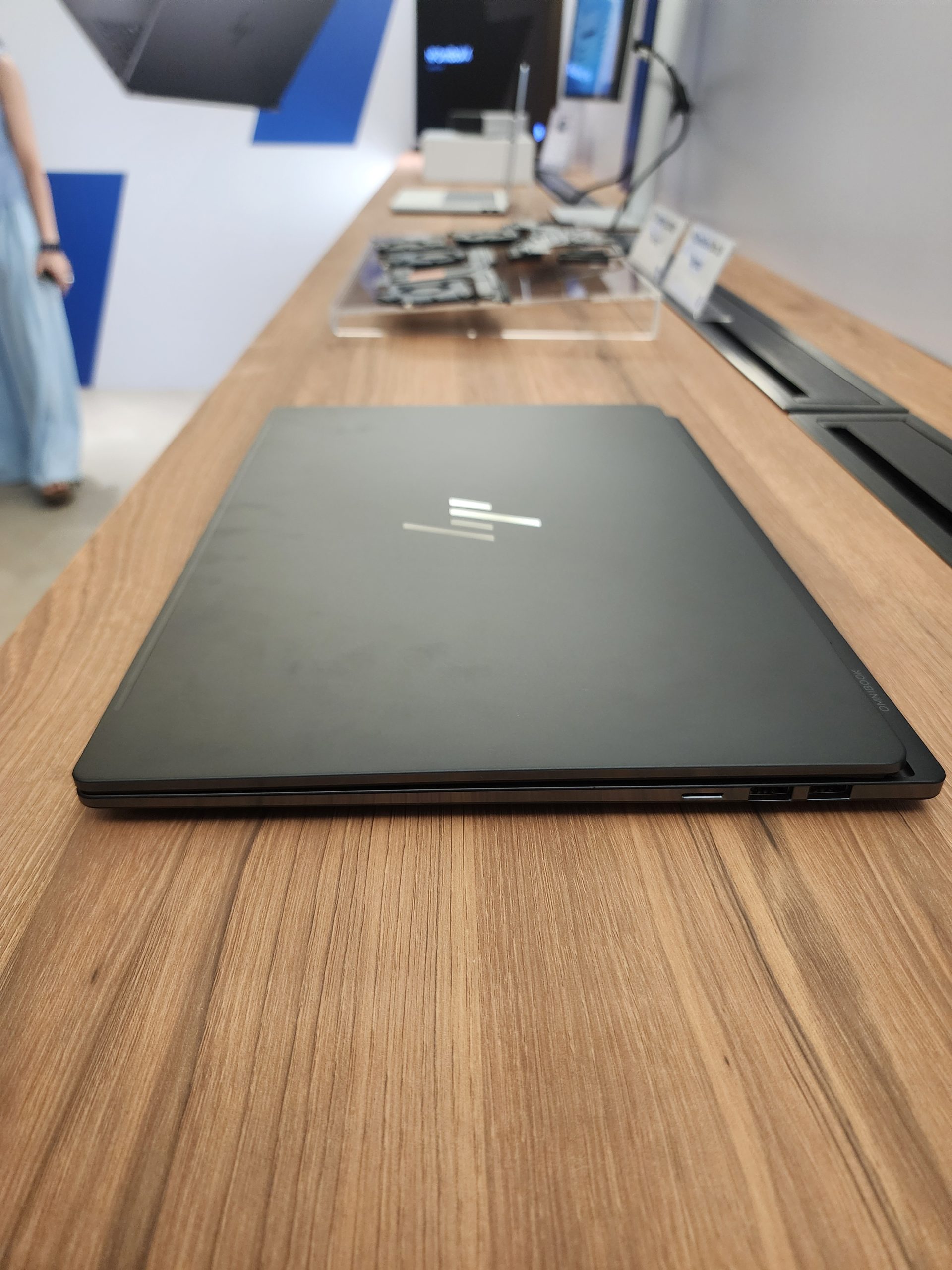 An HP laptop with a sleek, closed design is displayed on a wooden table, with other tech products blurred in the background.