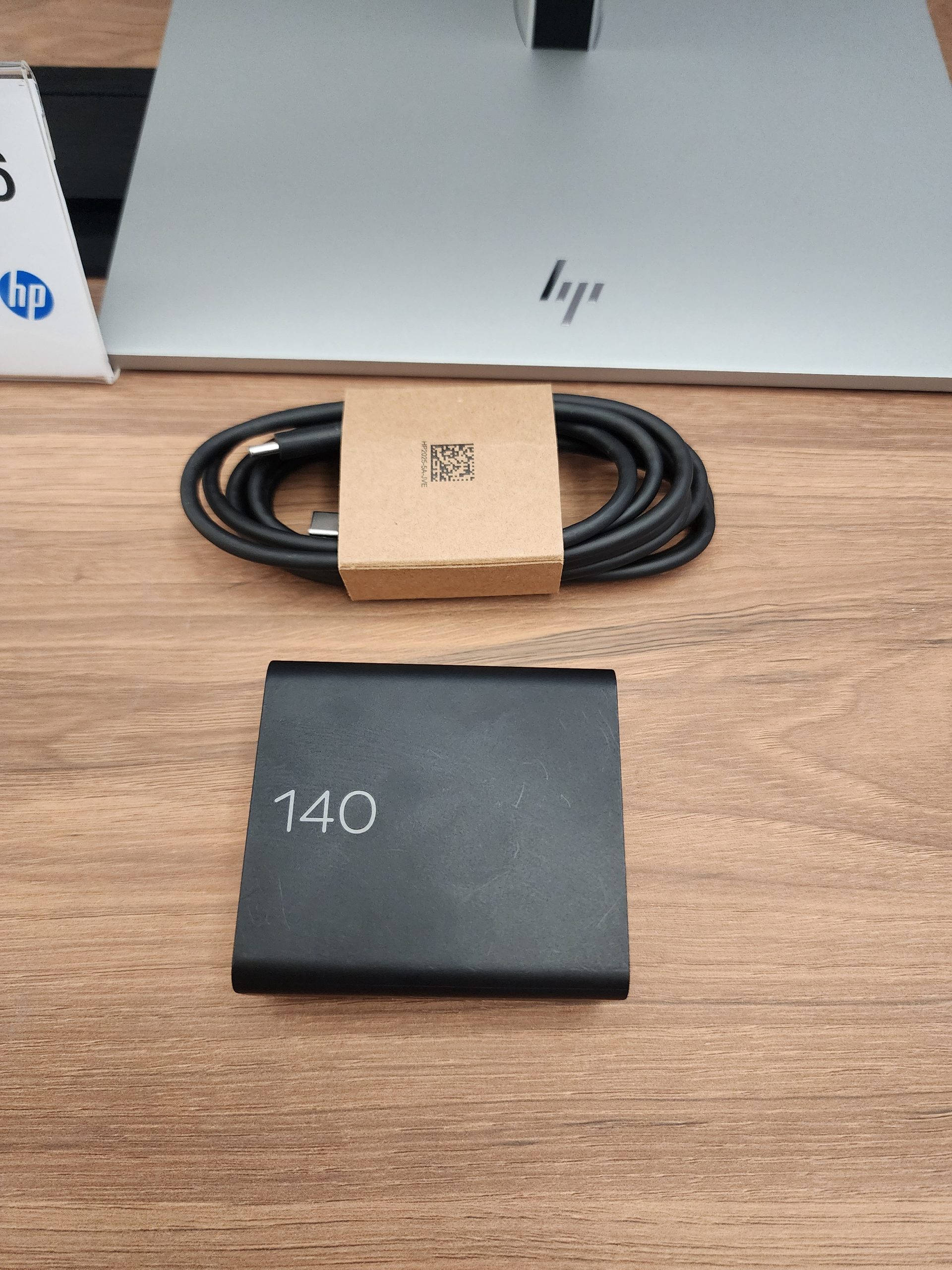 An HP power adapter labeled '140' is placed on a wooden table alongside a coiled charging cable, with an HP monitor stand visible in the background.