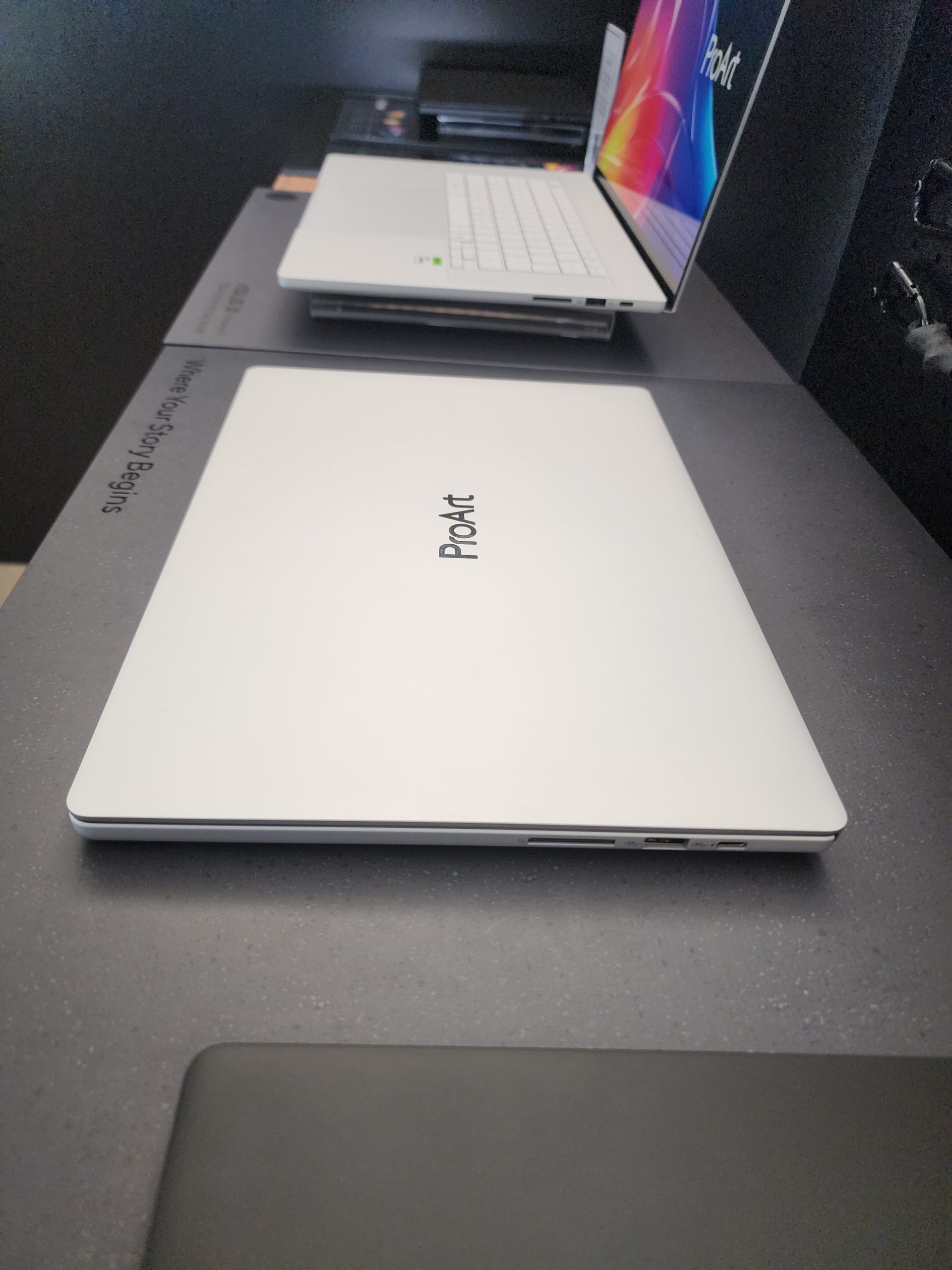 A lineup of ASUS ProArt laptops displayed with one screen showing a colorful wallpaper and the text 'Where Your Story Begins' printed on the surface beneath.